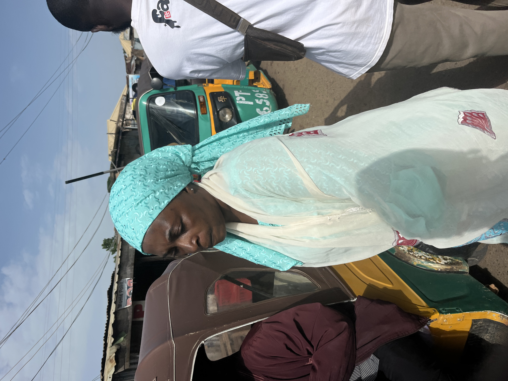 In Gura Top, Nigeria, a woman wearing a light blue headscarf and a flowing white garment stands beside a brown and yellow auto-rickshaw. She is looking downwards. To her right, a man in a white t-shirt and khaki pants stands facing away from the camera. In the background, a green auto-rickshaw with the license plate "PT 6589" is visible, along with a row of corrugated metal-roofed structures and overhead wires, suggesting a busy street or market environment. The sky is clear and blue.