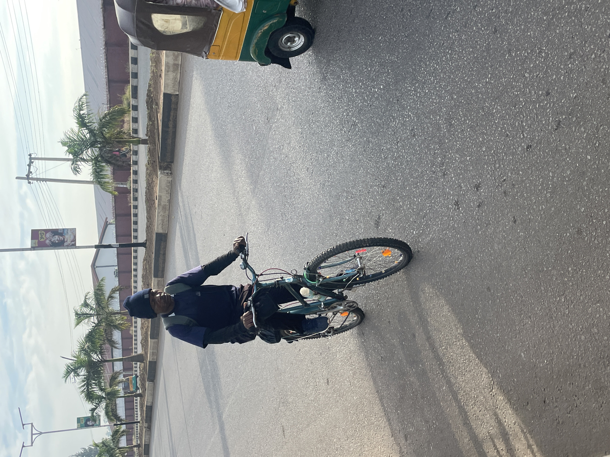 A man is riding a bicycle on a paved road in Jos, Nigeria. He is wearing a dark blue beanie, a dark blue shirt, and dark pants. The bicycle is dark green with a water bottle attached to the frame. In the background, there are palm trees, a building, utility poles, and a yellow and green autorickshaw. The sky is overcast, and the sun casts shadows on the road.