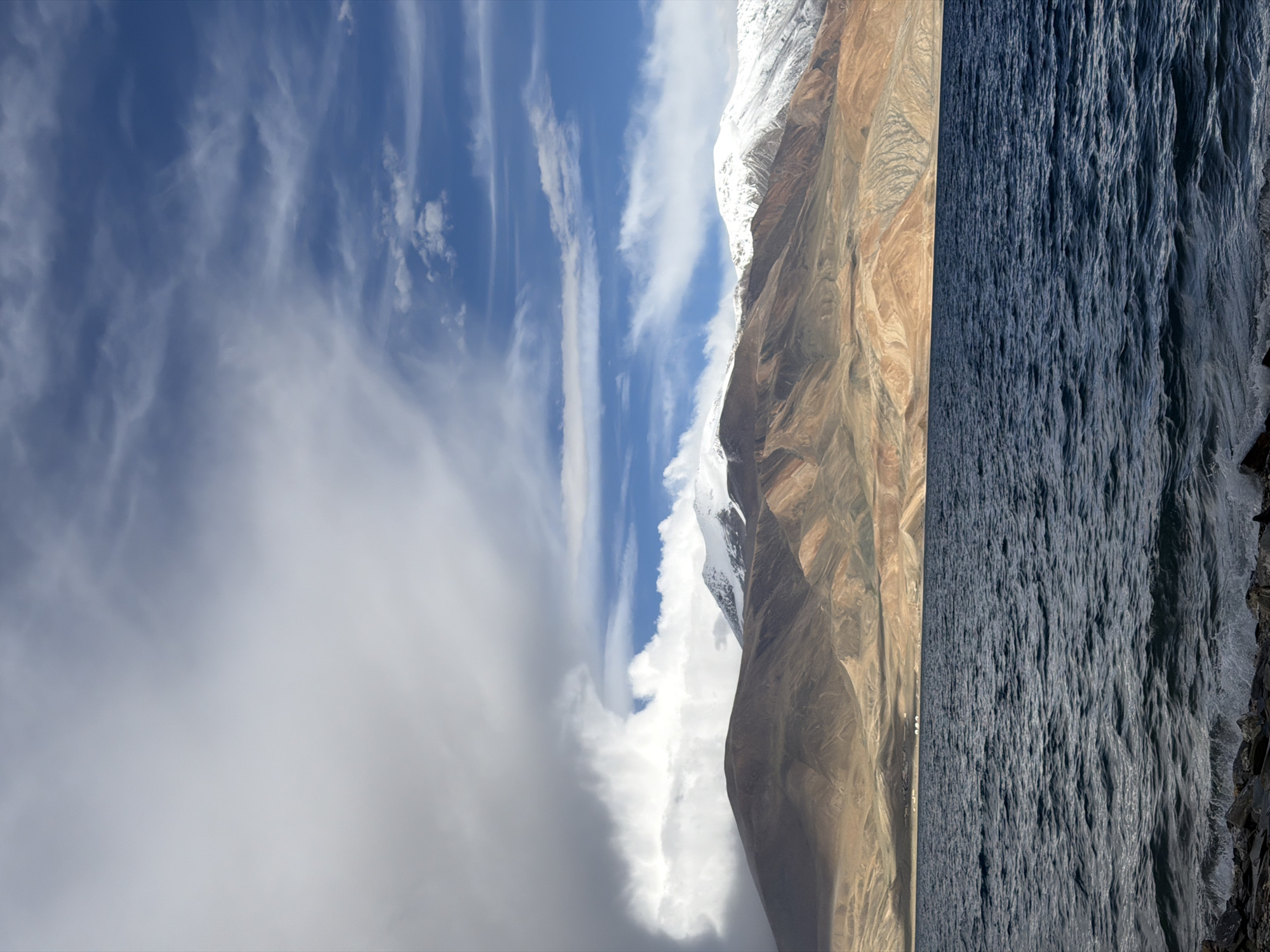 The image depicts a wide, dark blue body of water in the foreground, with ripples and waves indicating movement. Along the shore, dark rocks are visible. In the middle ground, a range of mountains rises, with the lower slopes appearing dry and barren, exhibiting shades of brown and ochre. The peaks of these mountains are capped with snow, creating a stark contrast with the earthy tones below. The sky above is a mixture of deep blue and large, dramatic clouds. Some clouds are thick and white, obscuring parts of the sky, while others are wispy and streaky, suggesting wind. There are no people or discernible man-made structures in the immediate scene, except for what appear to be a few small, white structures in the distance on the plains between the mountains and the water. The overall scene suggests a natural, possibly remote, mountainous landscape with a body of water.