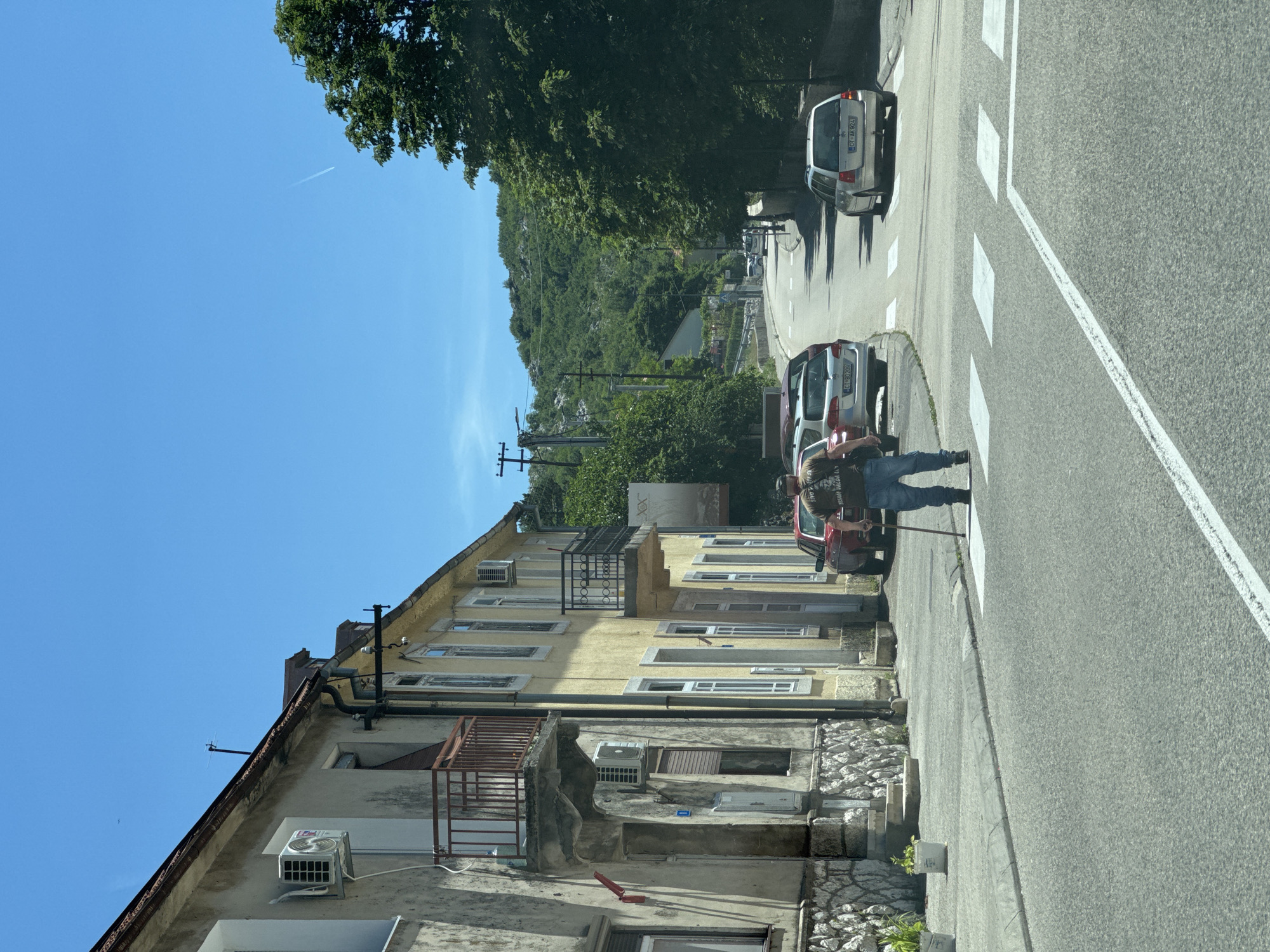 The image depicts a street scene in Cetinje, Montenegro, on a clear, sunny day. A man, appearing to be elderly and using a cane for support, is crossing a pedestrian crossing. He is wearing a t-shirt and blue jeans. Several cars are parked along the street and on the road, including a silver station wagon with the license plate CT AK924. On the left side of the street, there is a building with a stone foundation and a yellow-painted upper story. This building has several windows, balconies, and air conditioning units visible. Trees line the right side of the street, and a hilly, tree-covered landscape forms the background. The road is paved, and the sky is a vibrant blue with a faint contrail from an aircraft.