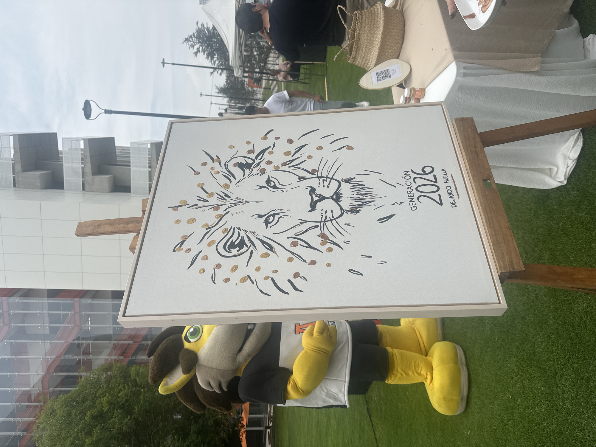 The image shows a drawing of a lion’s head in black lines with amber dots around the mane, which reads “Generación 2026 Dejando Huella." The drawing sits on a wooden easel. Below the artwork, part of a mascot costume, presumably for a lion, is visible. The mascot is wearing a white shirt with the letter “M” on it, black sleeves, and yellow gloves and boots. A manicured green lawn surrounds the easel and mascot. In the background, a building with a glass and red facade can be seen, along with other indistinct structures and people under a cloudy sky. The location is San Bernardino Tlaxcalancingo, Mexico.