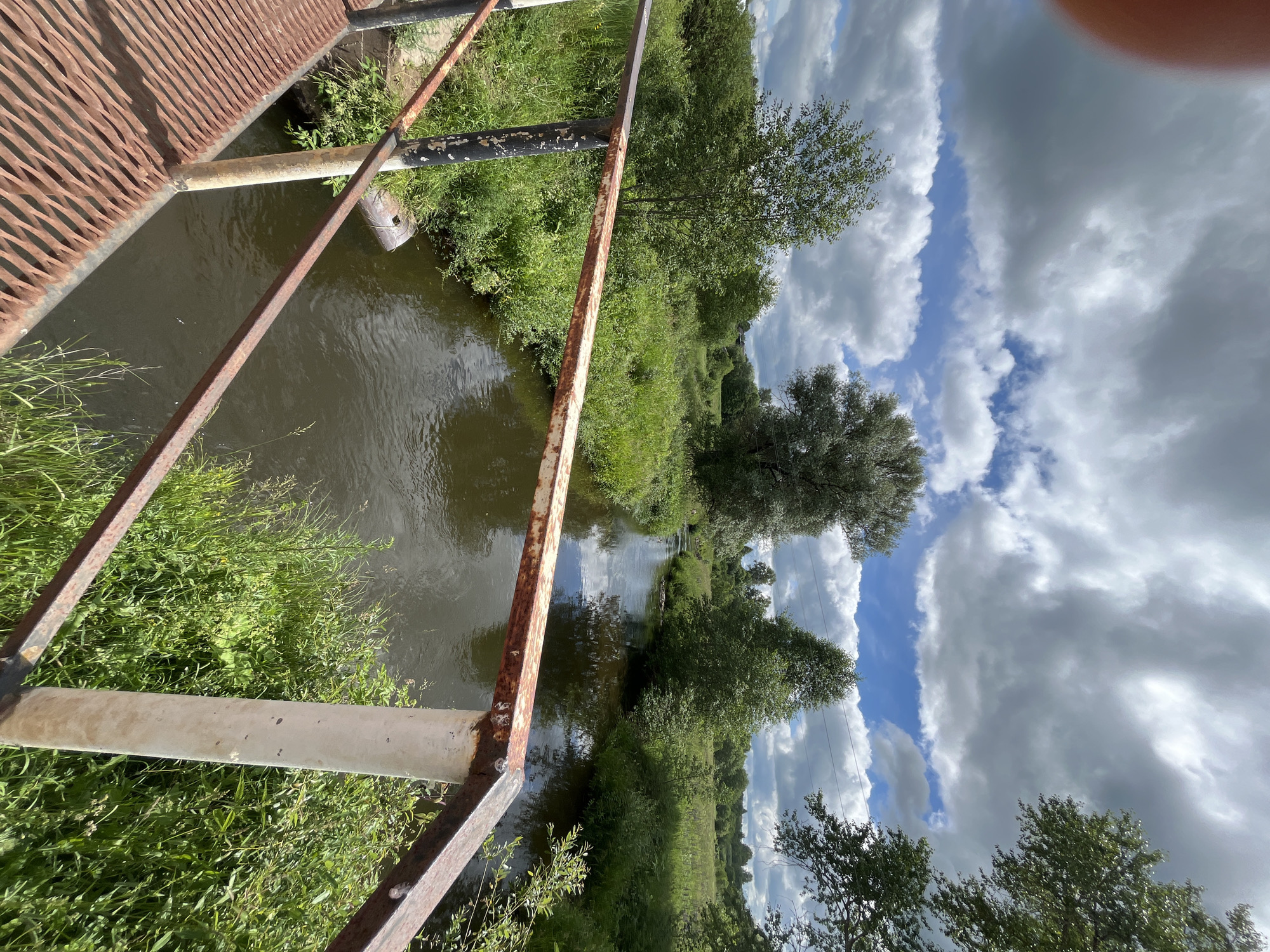 A rusted metal bridge railing overlooks a narrow, dark water river flowing through a lush green landscape under a partially cloudy sky. The riverbanks are densely overgrown with various types of green foliage and small trees. In the distance, more trees and a field are visible under the expansive sky filled with fluffy white and gray clouds. The scene appears to be in a natural, rural setting. No people or specific landmarks in the city of Уральское, Russia are identifiable in this image.