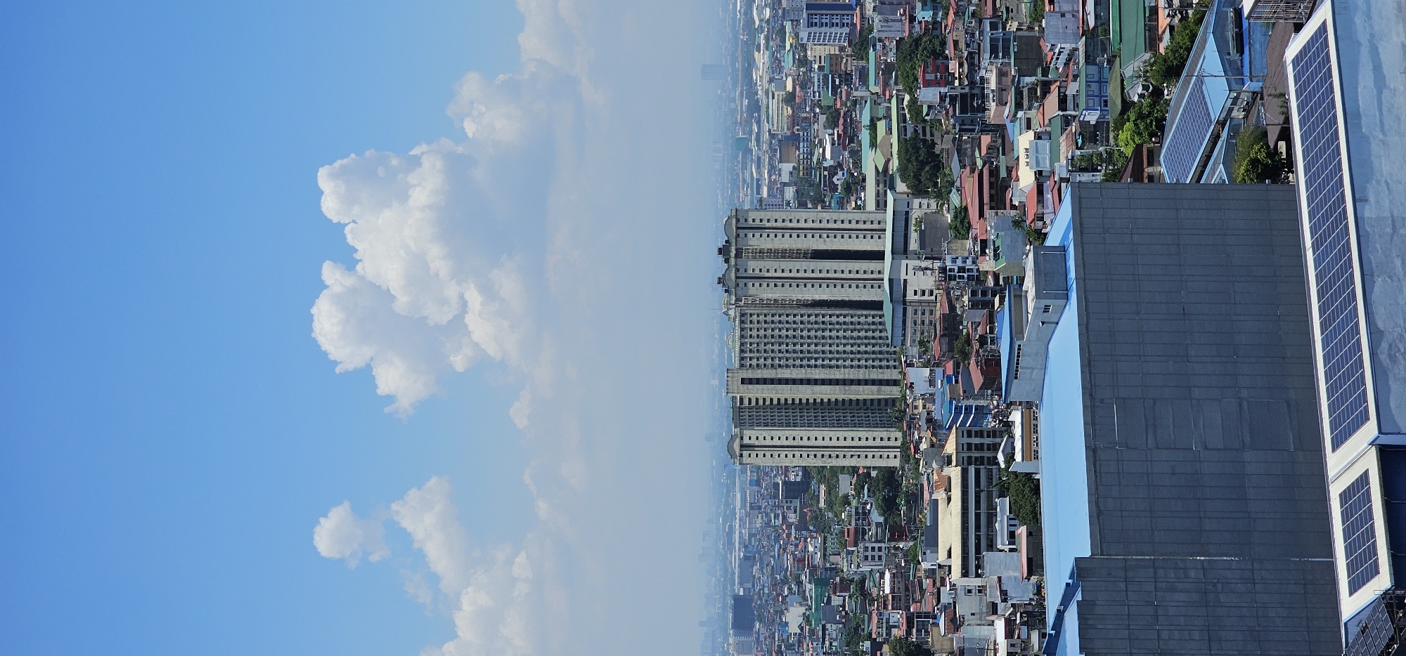 This is a vertical, eye-level, daytime, aerial view of a dense urban landscape in Makati City, Philippines. The image is dominated by a clear blue sky with scattered, fluffy white clouds. In the middle ground, two identical, tall, beige apartment buildings with repetitive dark-windowed facades stand prominently. Surrounding these central buildings is a sprawling cityscape of varied structures, including smaller residential buildings with colorful roofs, low-rise commercial buildings, and scattered trees. In the foreground, a large, modern building with a flat, light-blue roof is partially visible, with several rows of solar panels on its roof. The overall impression is of a busy, developed urban environment under a bright sky.