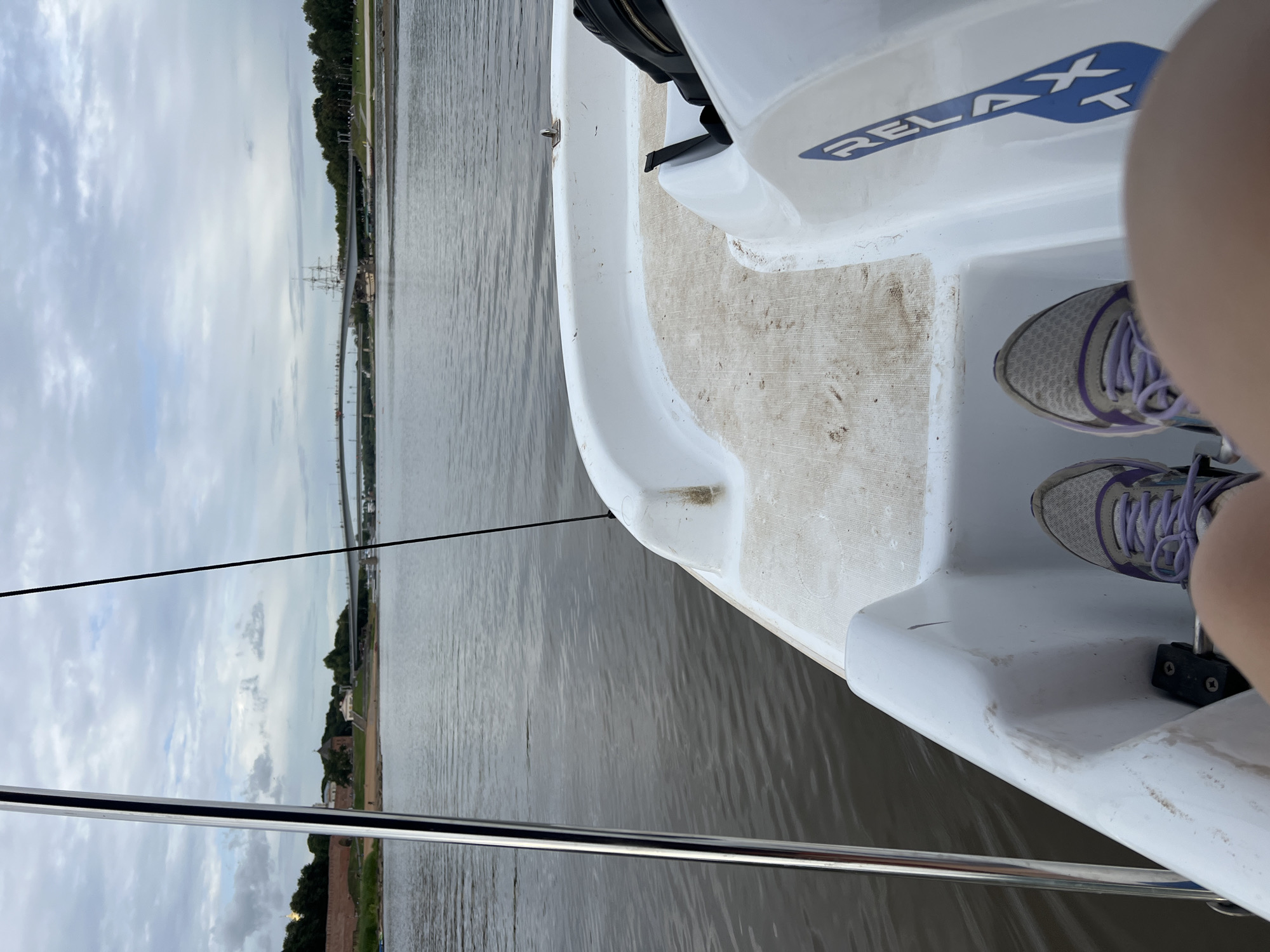 This is a first-person perspective photo taken from a boat on a body of water. The boat is white and appears to be a small recreational vessel. Visible on the boat is a blue decal with white lettering that reads "RELAX". There are two pairs of athletic shoes visible, one pair in the foreground and another further back, suggesting the presence of at least one person on the boat. The water is calm, reflecting the cloudy sky. In the background, the shoreline is visible with trees, buildings, and what appears to be a bridge or highway overpass. The sky is overcast, indicating a cloudy day. The scene is likely taking place during the daytime. The overall mood is relaxed and peaceful, consistent with the "RELAX" decal on the boat.