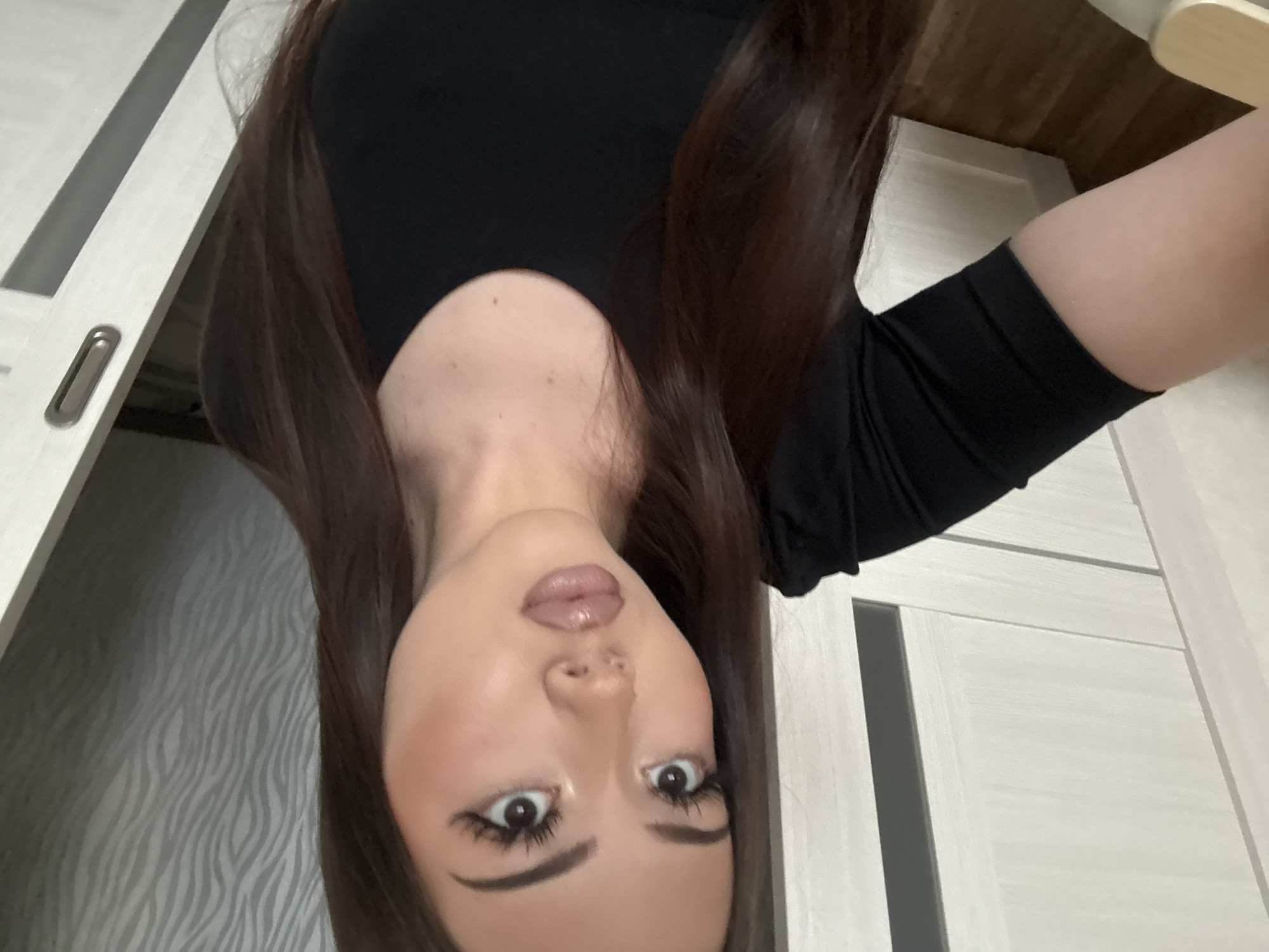 A woman with long brown hair is shown in the image, wearing a black long-sleeved top. The location appears to be indoors, possibly a residential space. The background shows a white door with gray accents and a wall with a wavy striped pattern. There is no definitive information to place the image in Naberezhnye Chelny, Russia.