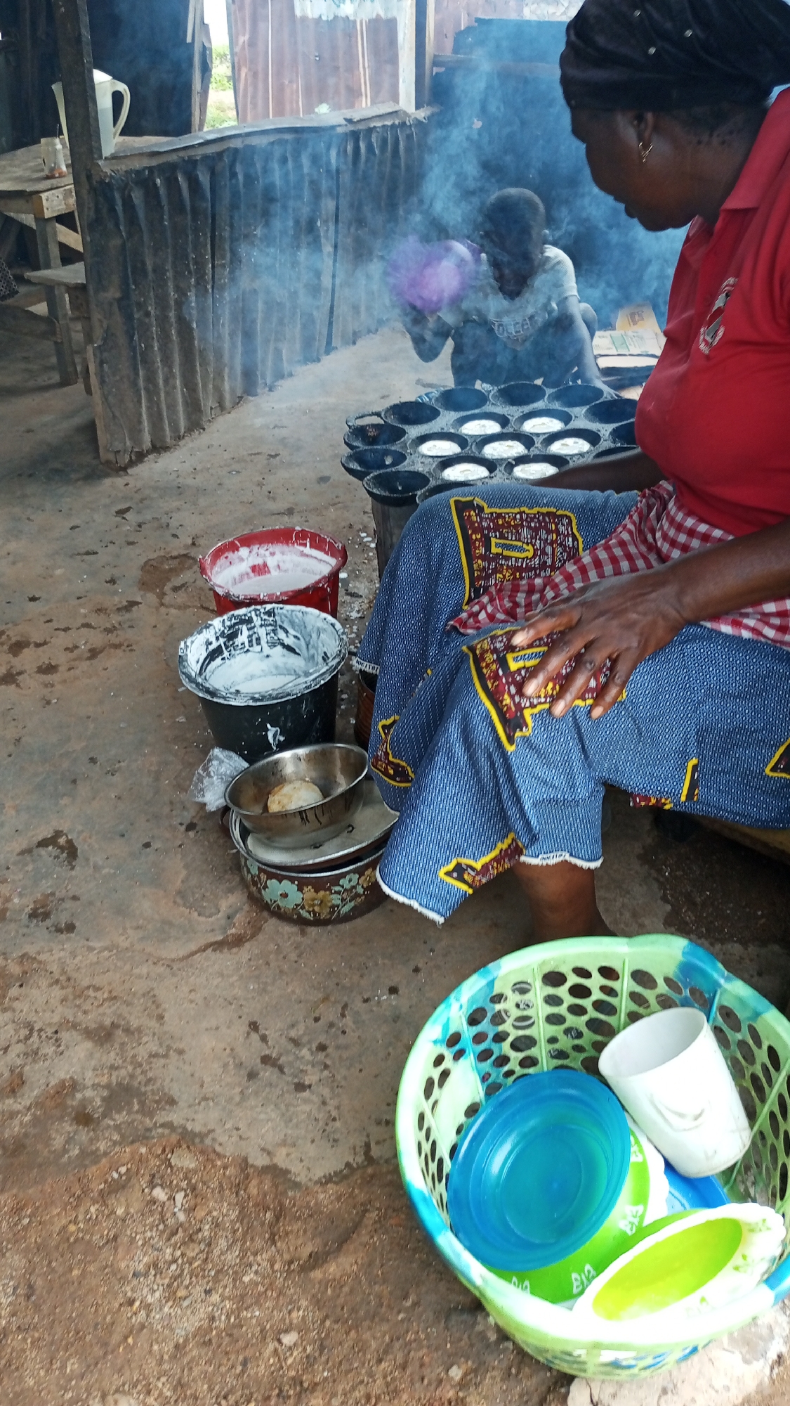 In Barikin Sabon Gidan Kanat, Nigeria, a woman in a red shirt and blue patterned skirt is cooking over a fire, with smoke rising around her. She is sitting next to several buckets, one red and one black, both filled with white batter. In front of her is a metal cooking apparatus with multiple round indentations, several of which contain what appear to be cooked pastries. She is holding a red and white checkered cloth in her hands. To her left, a child in a light-colored shirt is also near the fire, holding a purple object. The environment appears to be an outdoor cooking area, with a corrugated metal structure in the background and a dirt floor. To the right, a green plastic basket contains various plastic containers, including a blue bowl and a white cup.