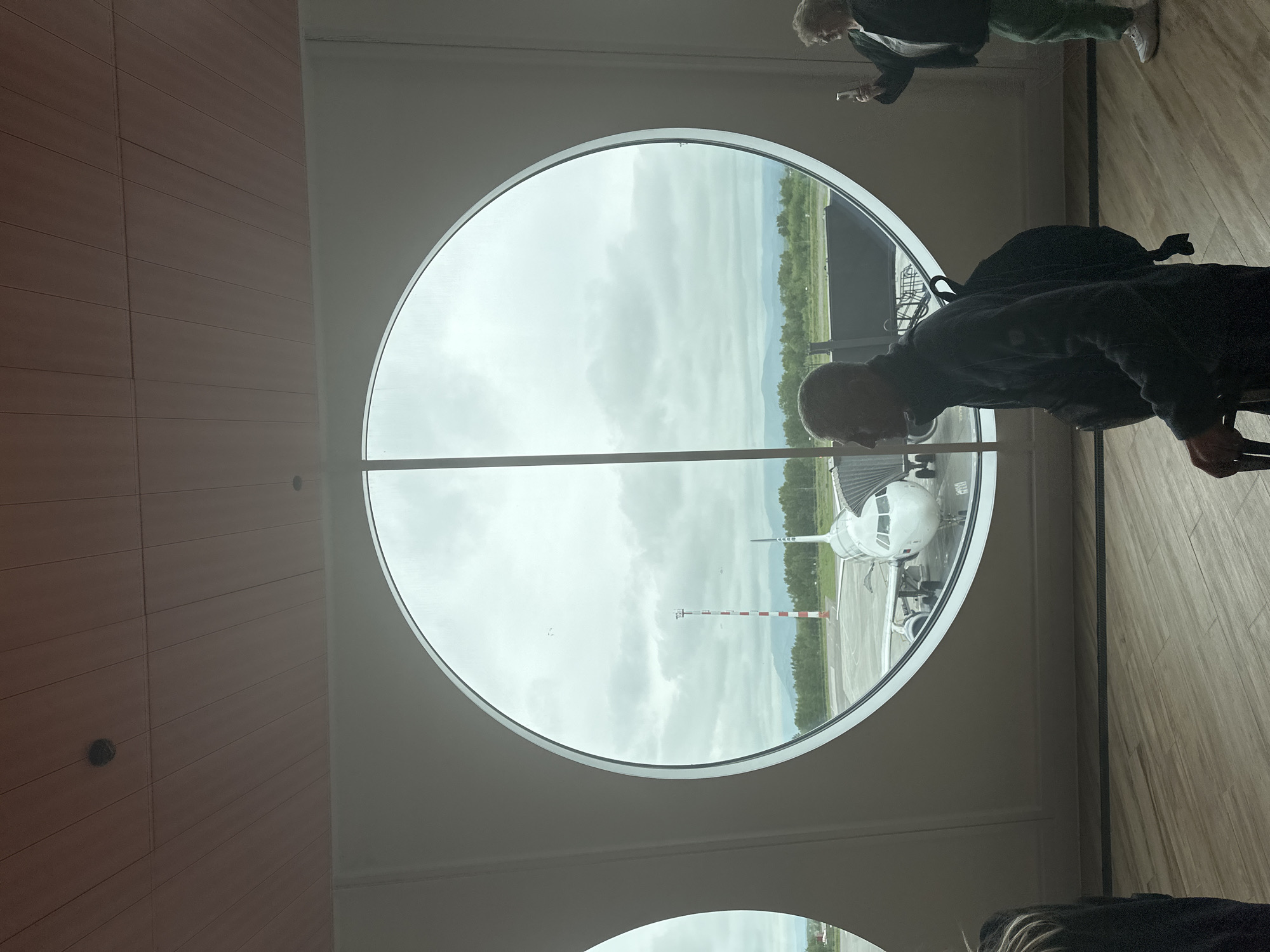 A man walks past a large circular window in what appears to be an airport terminal. The window overlooks a tarmac where a white airplane is parked, attached to a jet bridge. A red and white striped tower is visible in the background, along with trees and a cloudy sky. Another person, a woman, stands to the right of the window, holding a smartphone and taking a photo. The man in the foreground is wearing a dark jacket and backpack and is pulling a suitcase. The floor is made of light-colored wood planks. The ceiling is paneled with wood. The scene is located in Yuzhnyye Koryaki, Russia.
