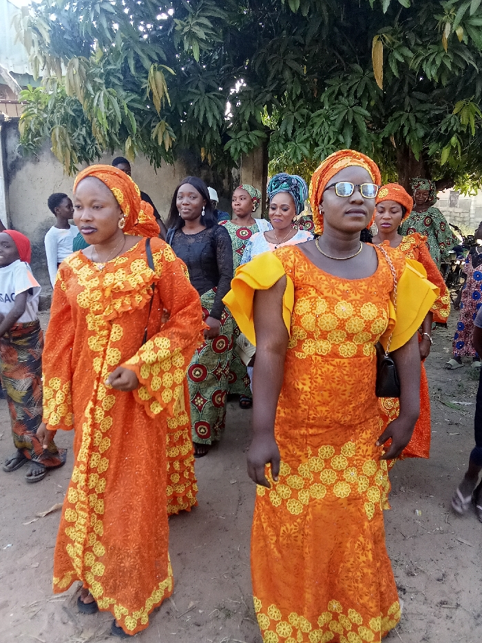 This image captures a group of people, primarily women, walking outdoors in what appears to be a communal gathering or event in Doma, Nigeria. The scene is set under the partial shade of large, leafy trees, possibly mango trees, with a dirt path underfoot and a light-colored wall visible on the left.
In the foreground, two women are prominently featured, both dressed in matching orange outfits with intricate yellow floral embroidery and coordinating headwraps. The woman on the left wears a long-sleeved dress with ruffled detailing on the bodice and embroidered cuffs, looking slightly to her left with a relaxed expression. The woman on the right wears a sleeveless dress with yellow ruffled shoulder sleeves, a gold-colored necklace, glasses, and carries a small black handbag. She gazes directly forward.
Behind these two, several other women are visible, also adorned in colorful traditional attire and headwraps, suggesting a uniform or celebratory dress code for the occasion. One woman in the middle distance wears a black lace top and a vibrant patterned skirt, while another behind her wears a lighter top and a different patterned skirt. On the far left, a child in a white t-shirt with the text "SAC" printed on it can be seen, partially obscured.
The overall atmosphere suggests a daytime event, likely late afternoon, under clear skies. The coordinated traditional African attire indicates a cultural, social, or ceremonial event.