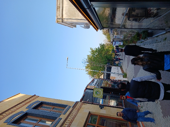 The image shows a narrow street in Istanbul, Türkiye, located at latitude 40.959 and longitude 28.823. The street is lined with buildings on both sides, with a yellow building on the left side. The street is paved with brick and is relatively empty. There are a few people walking down the street. One man is walking towards the camera on the left side of the street. A woman is walking in the opposite direction, closer to the camera. Three people are walking away from the camera, two men and a woman. The street is illuminated by a streetlamp and the sun. There is a sign for a restaurant or shop on the left side of the street.