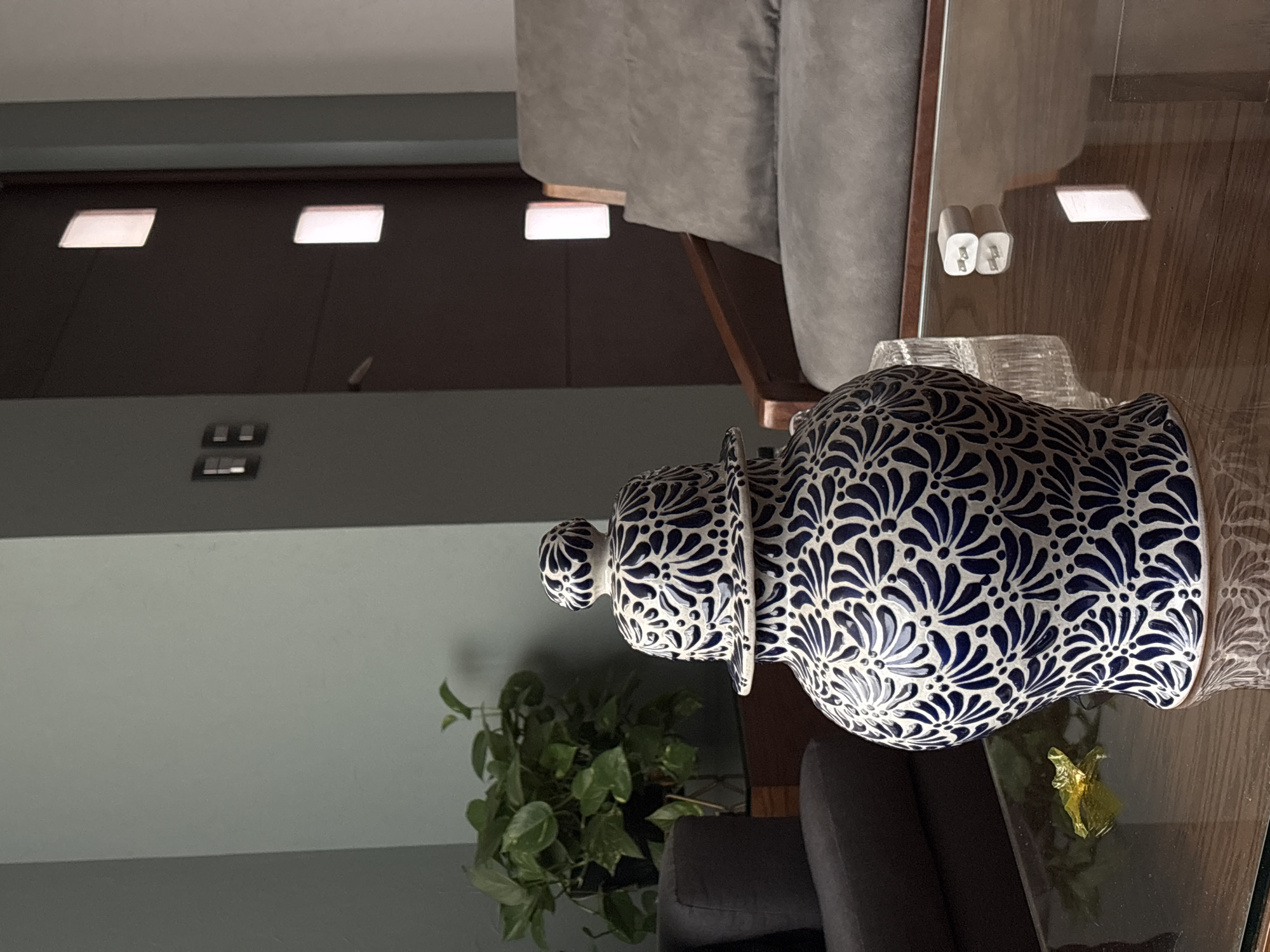 This is an interior photograph capturing a well-arranged living space, likely a living room, with a focus on a large, decorative ceramic vase.
The central subject is an ornate, lidded ceramic jar, predominantly white, adorned with intricate, symmetrical dark blue floral or stylized leaf patterns that cover its entire surface. This elegant piece rests on a rectangular coffee table, which features a transparent glass top reflecting overhead lights and other objects.
In the immediate vicinity of the vase on the table, a clear, textured glass item, possibly a holder or tray, is visible. A small, crumpled piece of gold-colored foil also lies on the glass surface. Part of a dark grey upholstered sofa or chair is seen in the lower right section of the frame, providing a contrasting texture.
In the background, a larger, light grey upholstered sofa with wooden armrest accents occupies the upper left portion of the image. Behind this sofa, a dark wooden cabinet or wall paneling spans across the mid-background. Above this dark surface, two rectangular recessed lights are set into a lighter-colored ceiling or upper wall, casting a soft glow.
To the left of the main vase, a vibrant green potted plant with numerous healthy leaves adds a natural element to the decor. The walls of the room appear to be a solid, neutral light grey or greenish-grey. Notably, two white power adapters, possibly phone chargers, are visible plugged into an outlet (or mounted on the wooden structure), and their distinct reflections with visible metal prongs are clearly seen on the glass tabletop.
The lighting in the room suggests either an evening setting or an interior primarily reliant on artificial illumination. There are no people present, and no specific activity is taking place. No discernible text is visible in the image. The overall impression is one of a meticulously designed and comfortable indoor environment.