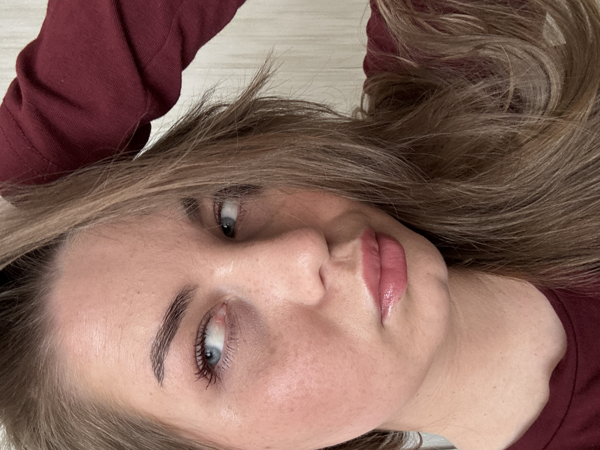 This diagonally oriented close-up image features a young woman looking directly into the camera. She has light blue eyes, defined eyebrows, and eyelashes enhanced with mascara. Her lips are full and adorned with a glossy, light pink or nude lipstick. Her complexion appears clear with a scattering of subtle freckles. She has long, light brown or ash-blonde hair that frames her face and spreads around her head. The woman is wearing a maroon long-sleeved top, parts of which are visible around her arms and shoulder. The background is a softly blurred, light-colored surface, possibly a wooden floor or a light-colored bed, indicating an indoor setting. Her expression is calm and neutral. No specific activity beyond posing for a photograph is discernible. There is no visible text in the image. While the image itself offers no explicit location cues, the provided context places the scene potentially in Chaikovsky, Russia. The time of day and weather cannot be determined from the visual information.