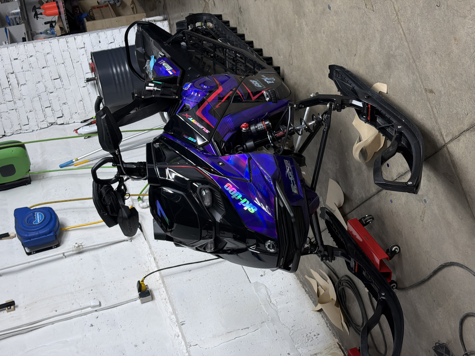 The image shows a black and purple Ski-Doo snowmobile, possibly in a garage or workshop. The snowmobile has the word "Ski-Doo" on it and "Summit X" on the side. The surroundings consist of tools, cords, and other equipment, suggesting a workspace. The location in Krasnoyarsk, Russia is impossible to confirm from the image itself.