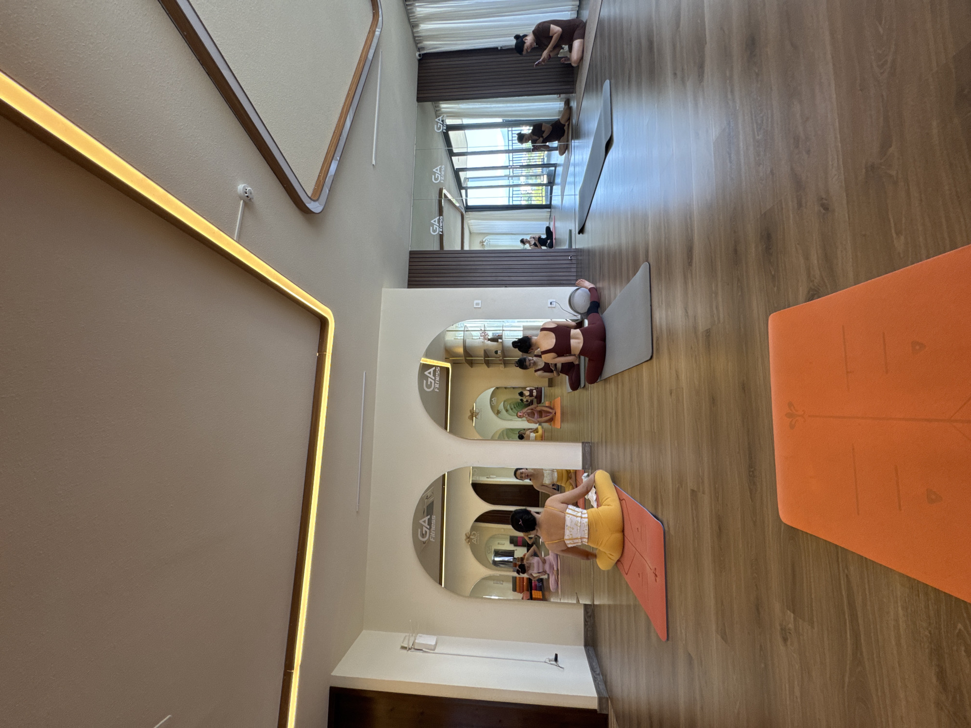 This image depicts the interior of a yoga studio, located in an unknown city. The studio features hardwood flooring. Several individuals are engaged in yoga poses on yoga mats, positioned on the floor. The walls are a light beige color. Large mirrors, framed by arched openings, reflect the yoga practitioners and studio space, creating a sense of depth and spaciousness. One person is wearing a yellow outfit and positioned on a peach-colored mat. An orange mat is placed near the right side of the image. A strip of light is on the wall.