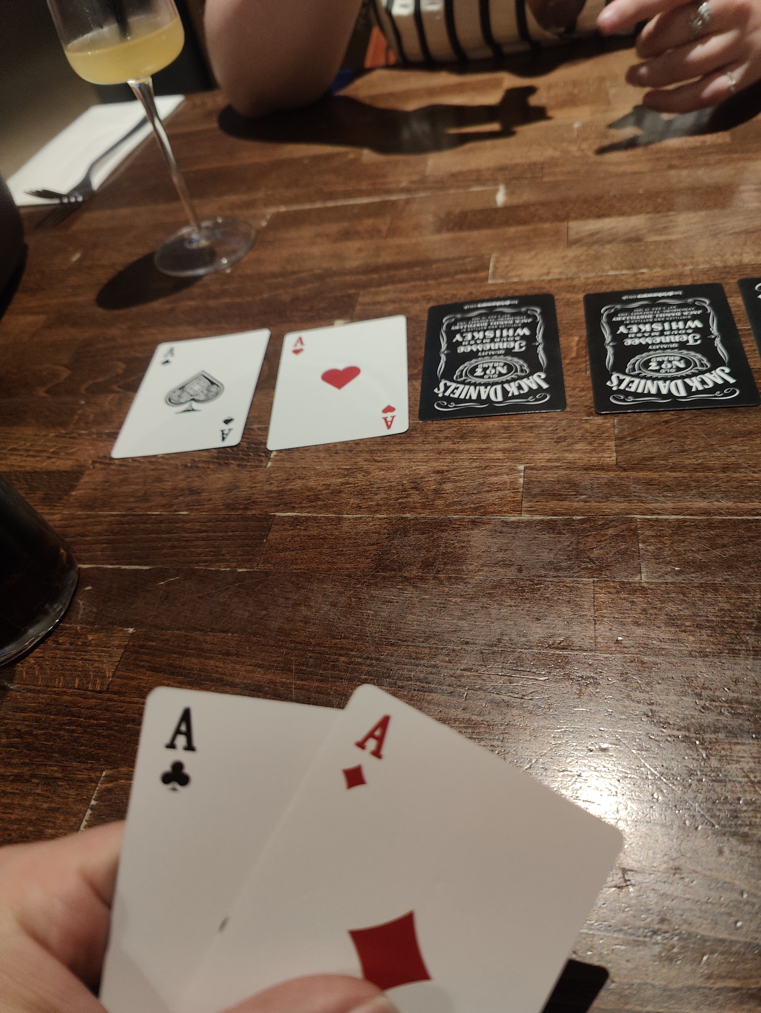 The image shows a close-up of a playing card game in progress on a wooden table. Five cards are visible, three face-up on the table and two held in a person's hand in the foreground. The table cards are: the Ace of Spades, the Ace of Hearts, and what appear to be two Jack Daniel's promotional cards (or potentially cards from a novelty deck). The person is holding the Ace of Clubs and the Ace of Diamonds. A glass of a yellow drink, a person wearing a striped shirt, silverware and a dark container are also visible in the background. The image does not provide any information to determine if the game is located in the city of East Suffolk, United Kingdom.