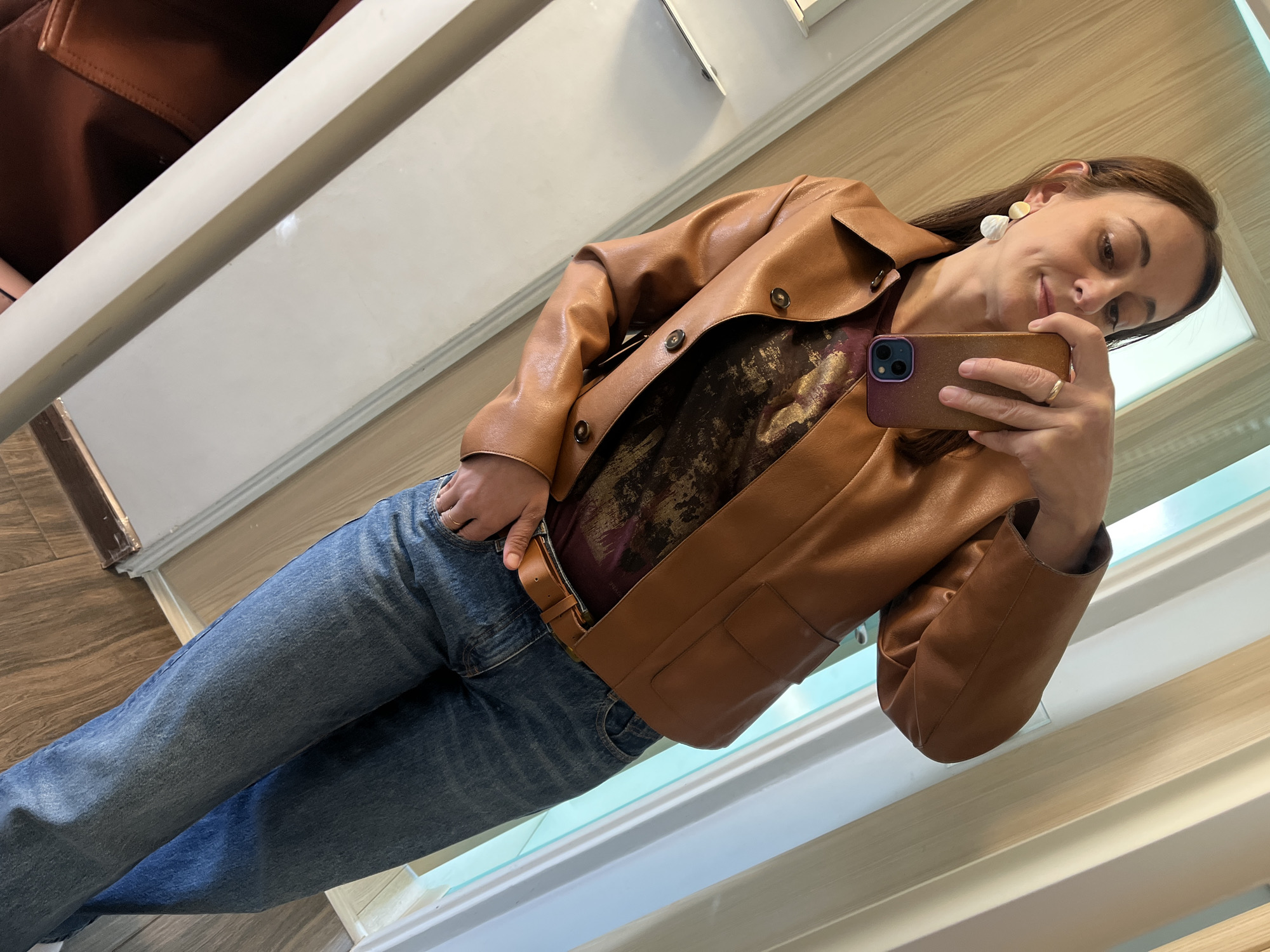 A young woman with brown hair, wearing a tan leather jacket over a dark patterned shirt, blue jeans, and a tan belt, stands and takes a mirror selfie. She has two white earrings on her left ear and a ring on her right hand. The background shows a hallway with light wood-paneled walls and a door.