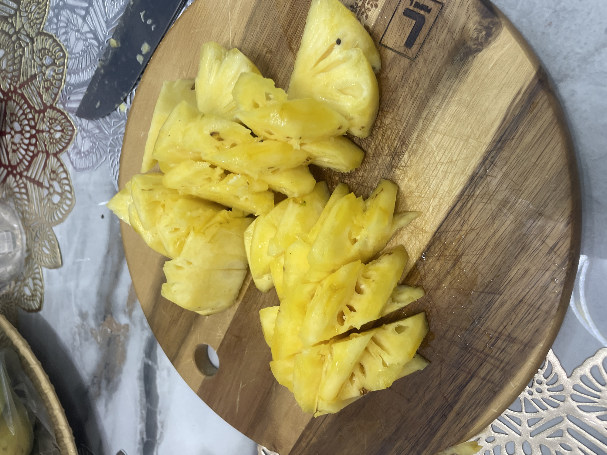 This media file shows sliced pineapple on a wooden cutting board. The pineapple pieces are bright yellow and appear juicy, with some pieces showing the fibrous core. The cutting board is round and made of wood with visible grain and scratches, suggesting it has been used frequently. To the left of the cutting board, a part of a knife with a dark handle is visible. The background is a patterned surface, possibly a tablecloth or placemat, with an intricate design in metallic gold and white. Further to the left and below, a portion of a woven basket is visible, hinting at a domestic or food preparation setting. The lighting suggests it is daytime, possibly indoors, as there are no strong shadows or direct sunlight. The overall scene conveys a sense of preparing food, likely for consumption or storage. There is no visible text in the media file. The location context is Nha Trang, Vietnam.