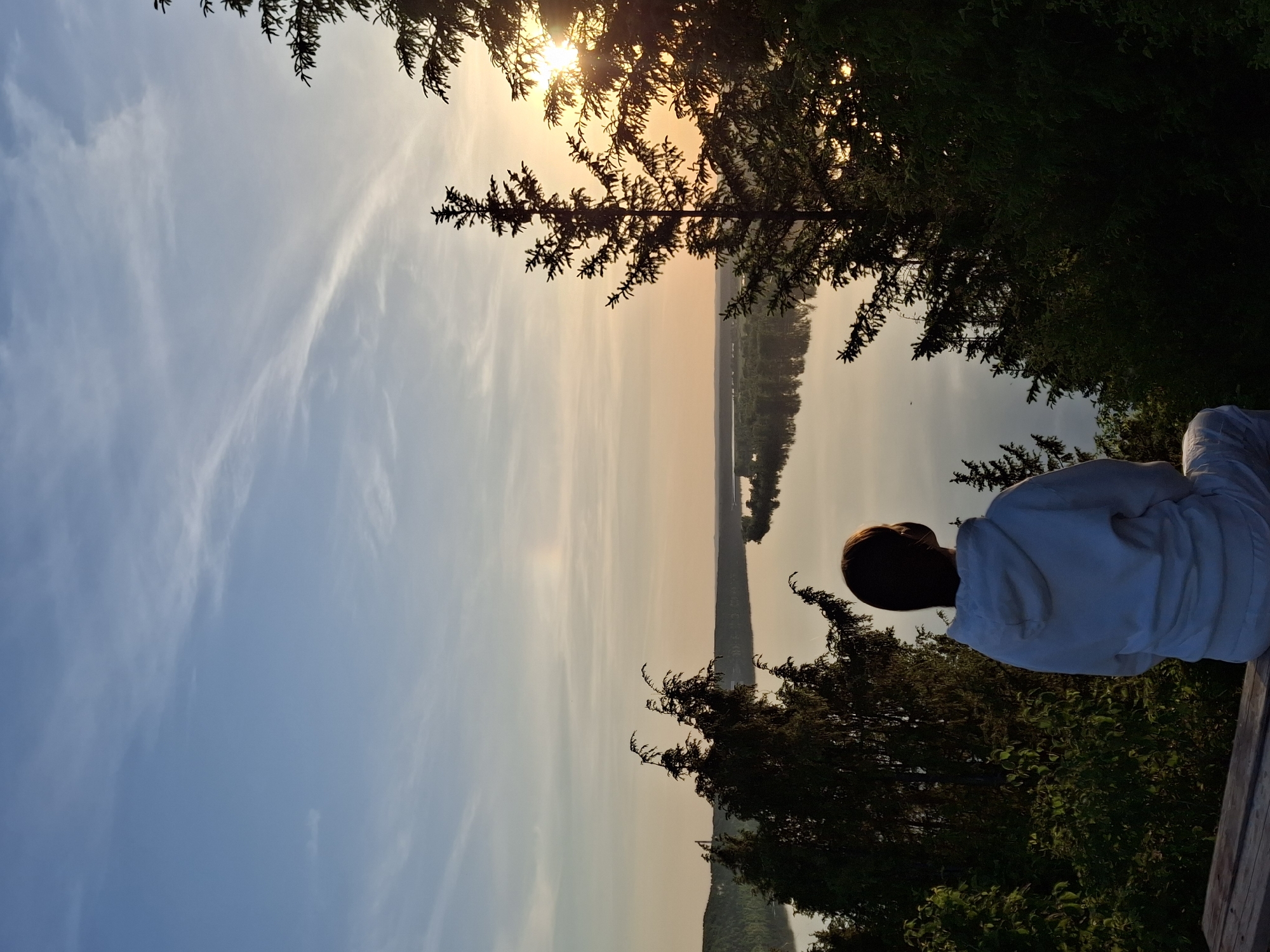 A person with short brown hair and a white sweatshirt sits facing a body of water from a wooden bench, framed by trees on either side. The sun is setting, casting a warm glow over the horizon in Прикамский, Russia. The sky is a gradient of blue to orange. A thin strip of land borders the water's edge, dotted with trees. The vegetation around the bench is lush and green.