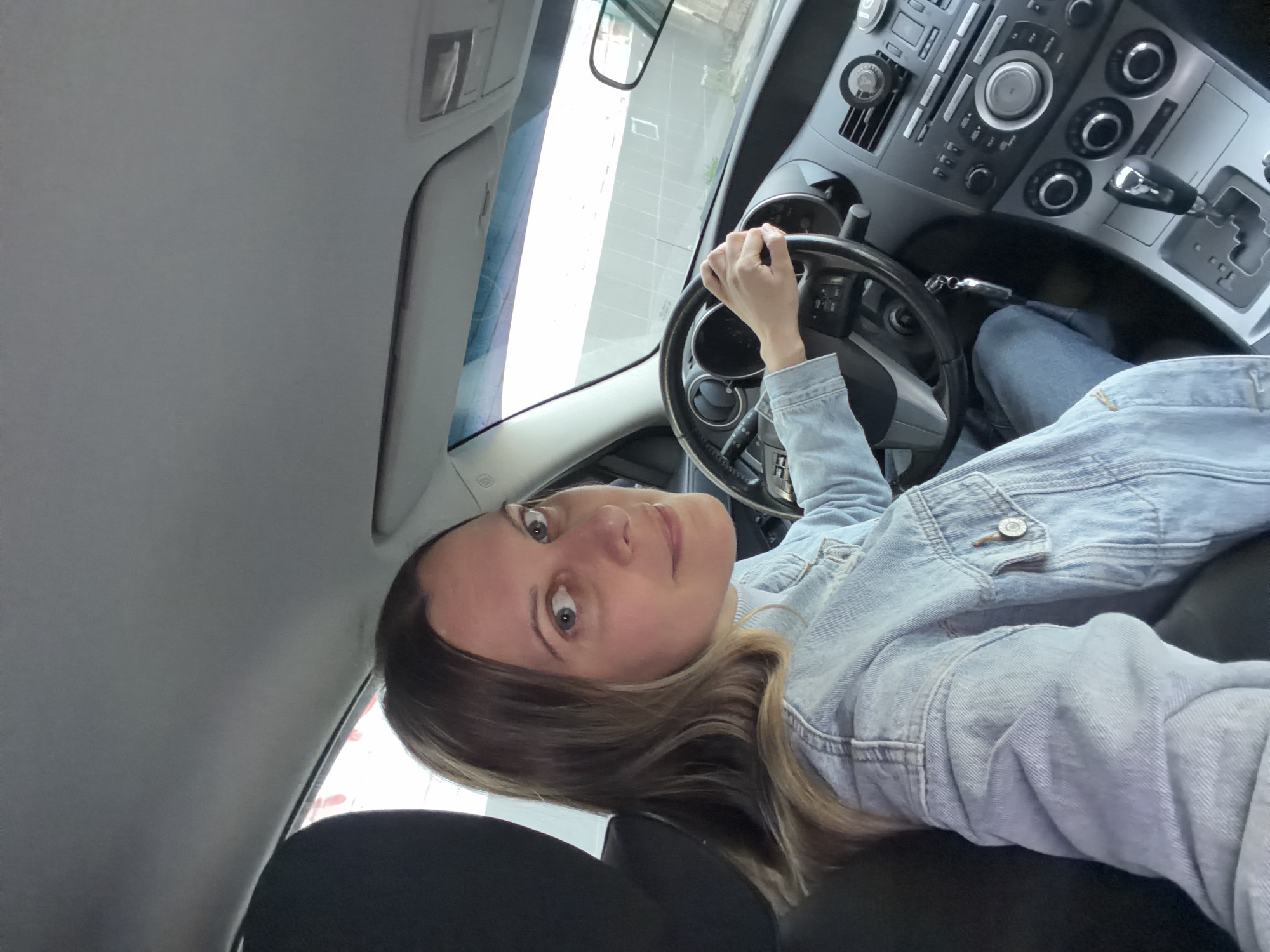 A woman with shoulder-length hair, wearing a light blue denim jacket and jeans, is seated in the driver's seat of a car. Her left hand is on the steering wheel. The car's dashboard is visible, as is part of the interior, including the gearshift and center console. The location is potentially within a city; though not certain, there is a very low chance it is in Izhevsk, Russia given the available visual information alone. There are no specific indicators connecting the image to that city.