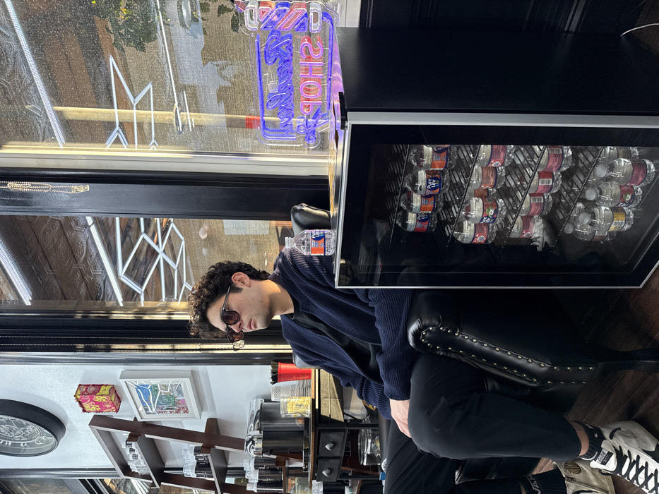 This image depicts the interior of a barbershop located in Boston, United States. The exact street address or specific neighborhood within Boston cannot be determined from the visual information provided.
A male individual with dark, curly hair, wearing dark sunglasses, a dark blue ribbed cardigan over a dark shirt, and black trousers, is seated in a black leather armchair with brass studding, facing towards a large window to his right. A bottle of Pure Life water rests on the armchair's right armrest. His left foot, clad in a black sock and a white and black high-top sneaker with a star emblem, is visible on the dark wood floor.
To the immediate right of the armchair, a compact black mini-fridge with a clear glass door is stocked with multiple rows of Pure Life bottled water. On top of the mini-fridge, a lit neon sign displays "BARBER SHOP" in blue and red.
In the background to the left, a dark wooden shelving unit holds various containers, likely haircare products. Below it, a dark wooden cabinet features a chrome dispenser, red plastic cups, and a clear container with yellow, possibly straw-like, items. The wall decor includes a large dark circular clock, a decorative red and gold rectangular box, and a framed colorful landscape artwork. Bright natural light enters the space through the large window, reflecting interior elements such as a patterned ceiling and another neon sign. The exterior view through the window is indistinct.