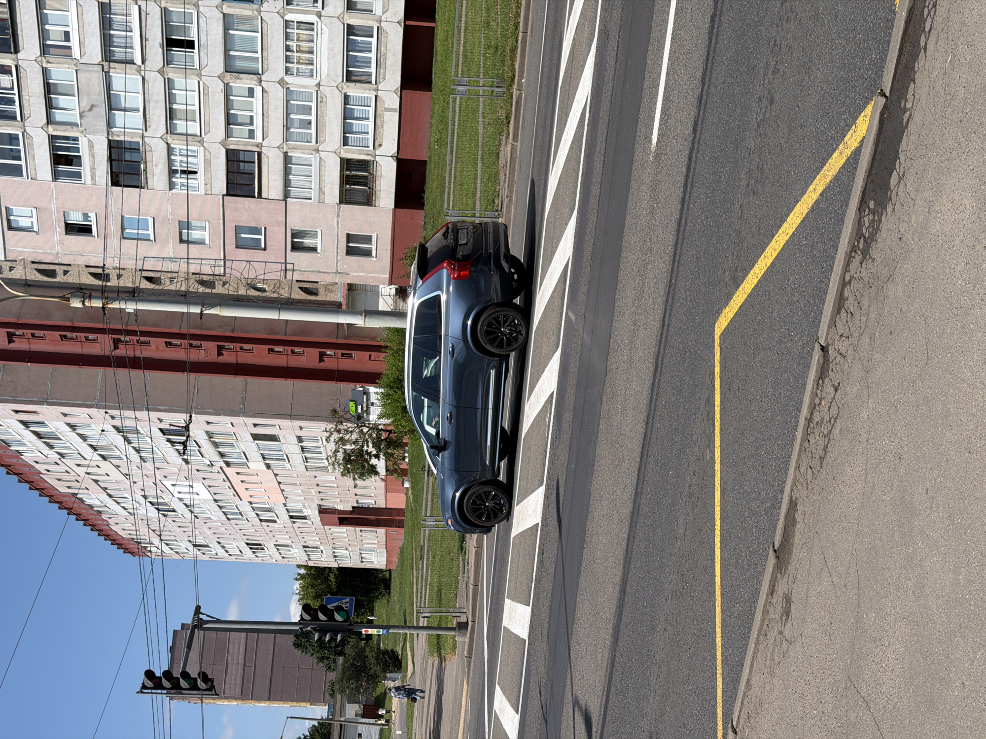 The image shows a street scene in Minsk, Belarus. In the foreground, a dark blue SUV is driving across a pedestrian crossing. The SUV has black rims and a white roof rack. The pedestrian crossing is marked with white diagonal stripes. The road is asphalt. In the background, there is a tall apartment building with multiple windows and balconies. The building is light pink and has a red trim at the top. To the left of the apartment building, there is a traffic light and a shorter building, possibly another apartment building. A few trees and a metal fence are also visible between the road and the buildings. The sky is blue with a few scattered clouds. A person is walking on the sidewalk in the distance. The overall scene appears to be a typical urban environment on a sunny day.