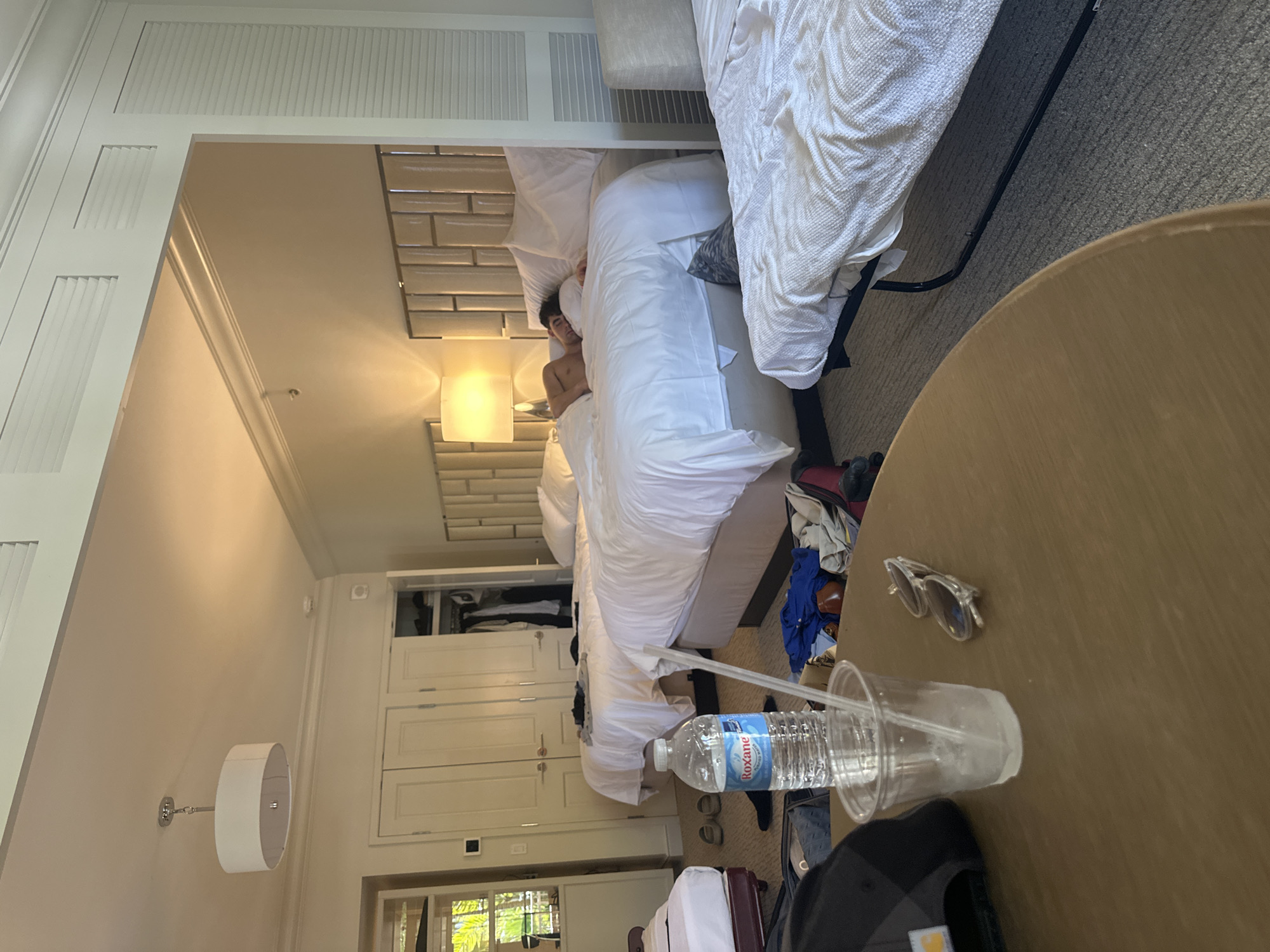The image shows a bedroom scene, possibly in a hotel in Aventura, Florida, United States. There is a person sleeping in one of two beds in the room. The room has light-colored walls, built-in closet space, a window, and a light fixture. On a table in the foreground, there's a plastic cup, a water bottle (with the Roxane label), sunglasses, and a baseball cap.