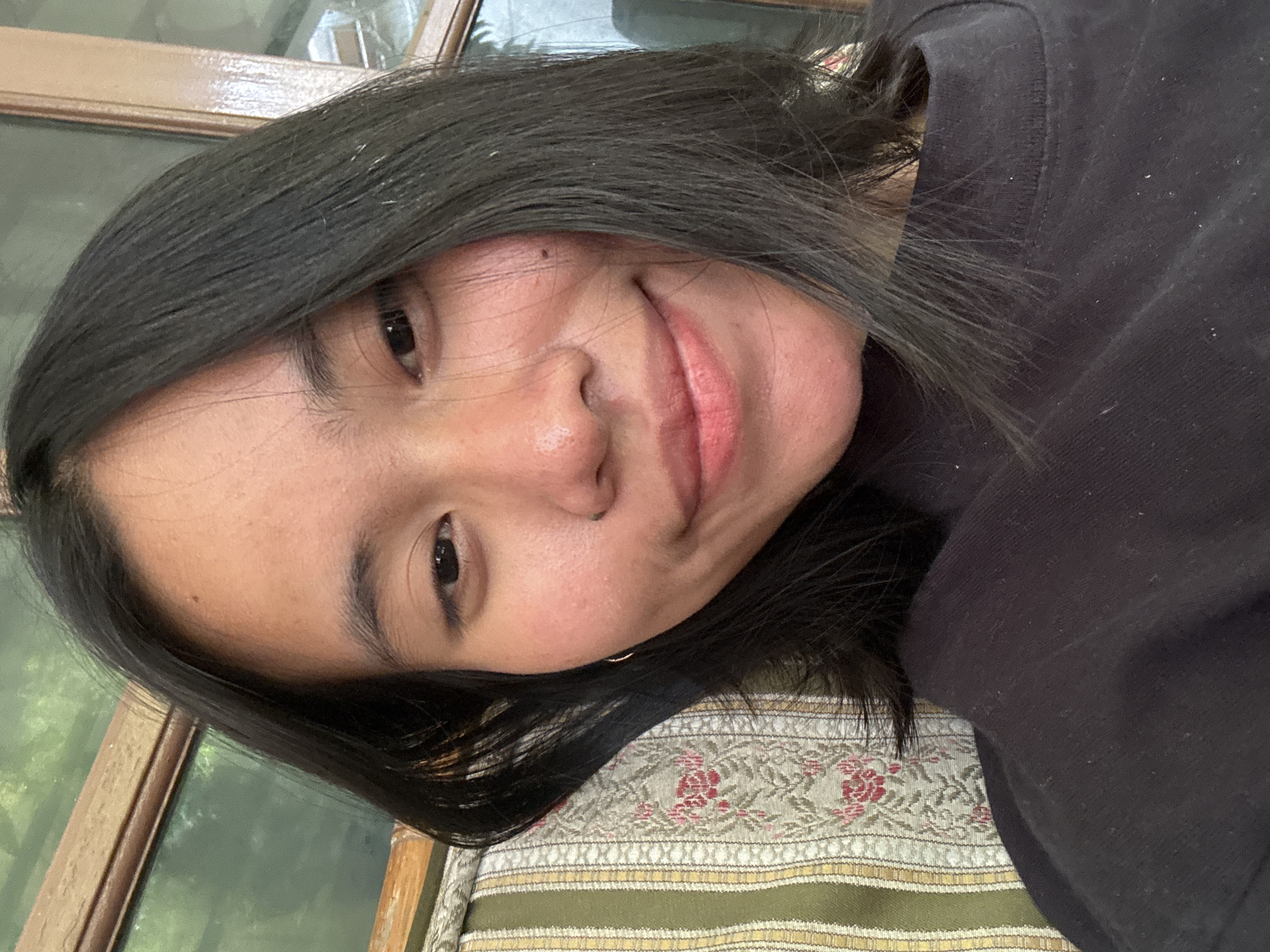 A medium shot selfie features a young woman with medium-length dark hair framing her face. She is looking directly at the camera with a subtle smile, her lips appearing slightly glossed. Her skin is clear with a few subtle blemishes visible. She is wearing a dark t-shirt. Behind her, a patterned cushion with green, yellow, and red floral designs is partially visible. A wooden window frame is also discernible in the background, with greenery beyond it, suggesting an outdoor or semi-outdoor setting. The overall lighting is soft and natural, indicating the photograph was taken during the daytime, likely indoors or in a shaded outdoor area. The presence of a patterned cushion and wooden window suggests a residential setting, possibly a home in a neighborhood within South Jakarta, Indonesia.