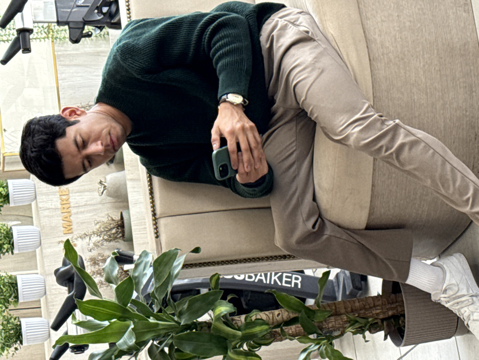 A man in a green sweater and khaki pants reclines on a beige sofa, gazing down at a green phone in his hand. He wears a gold watch. A potted plant with long, green leaves sits near the sofa's base. In the background, there are several small white potted plants on a wall, with the word "MARKET" visible. Part of an exercise bike is also present in the image.