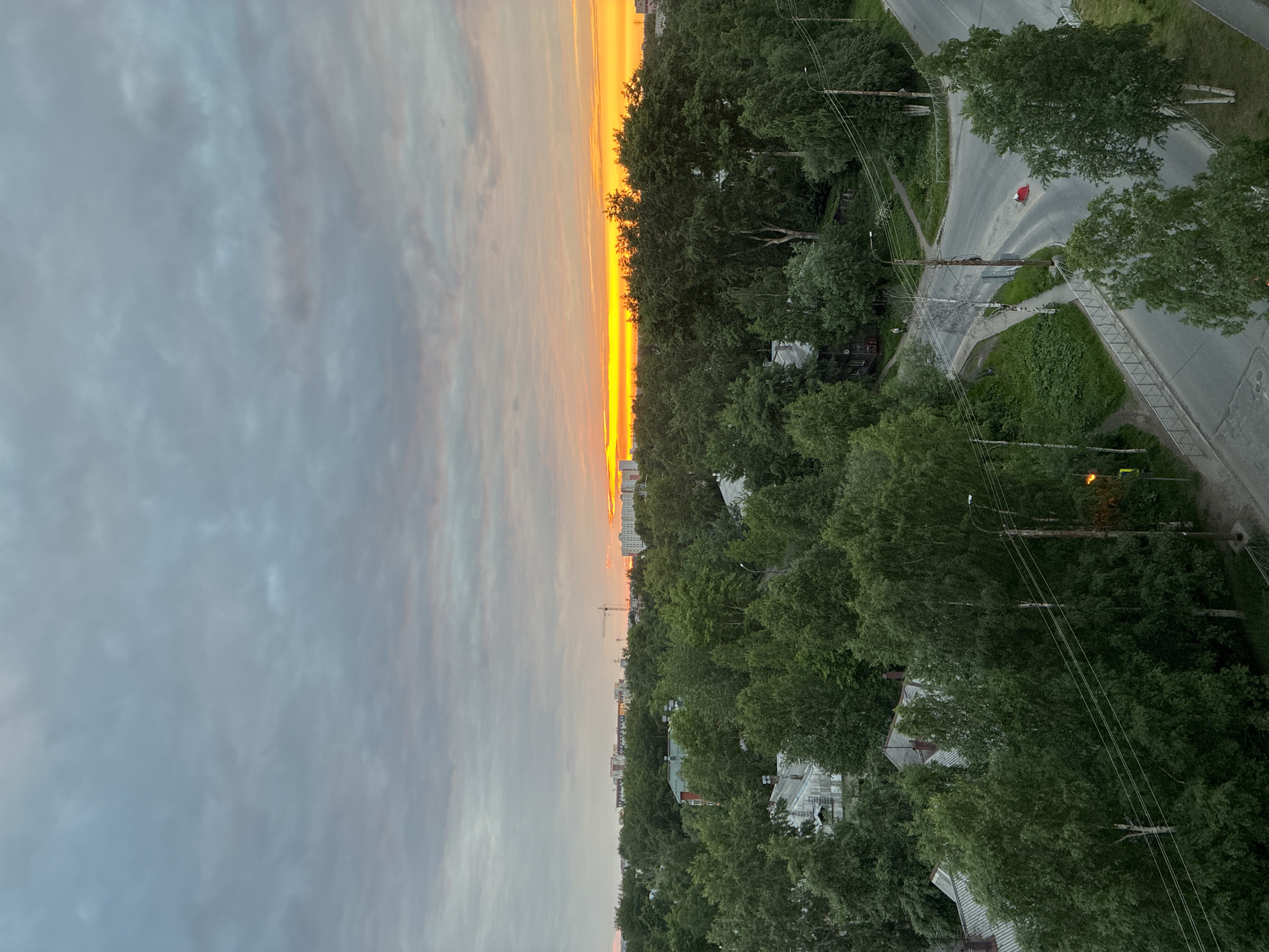 An aerial or high-angle photograph depicts a verdant urban landscape at either sunrise or sunset. The sky, occupying the upper portion of the image, is predominantly overcast with mottled grey clouds that transition to a brilliant band of intense orange and yellow light along the horizon, particularly prominent on the right.
In the background, on the horizon line and partially obscured by trees, various buildings are visible, including taller, multi-story structures on the left and a large construction crane silhouetted against the bright sky in the center-right. Further right, additional buildings reflect the golden light.
The mid-ground is dominated by a dense canopy of lush, green deciduous trees, suggesting summer or late spring foliage. Interspersed among these trees are numerous residential buildings, some featuring older-style corrugated or shingled roofs, typically light grey or brown, and others with flatter, more modern designs.
The foreground focuses on a section of the urban environment. A paved asphalt road, light grey in color, curves and forms an intersection in the lower right. Sidewalks run parallel to the roads, bordered by metal railings. A prominent wooden utility pole, equipped with multiple overhead wires and a streetlight, stands at the intersection. Another streetlight to its left is illuminated with an orange glow. On the curved portion of the road, a distinct bright red object, possibly a bag or crumpled fabric, lies on the asphalt. In the upper left of the foreground, two small, indistinct figures are visible walking on a dirt path in the distance.