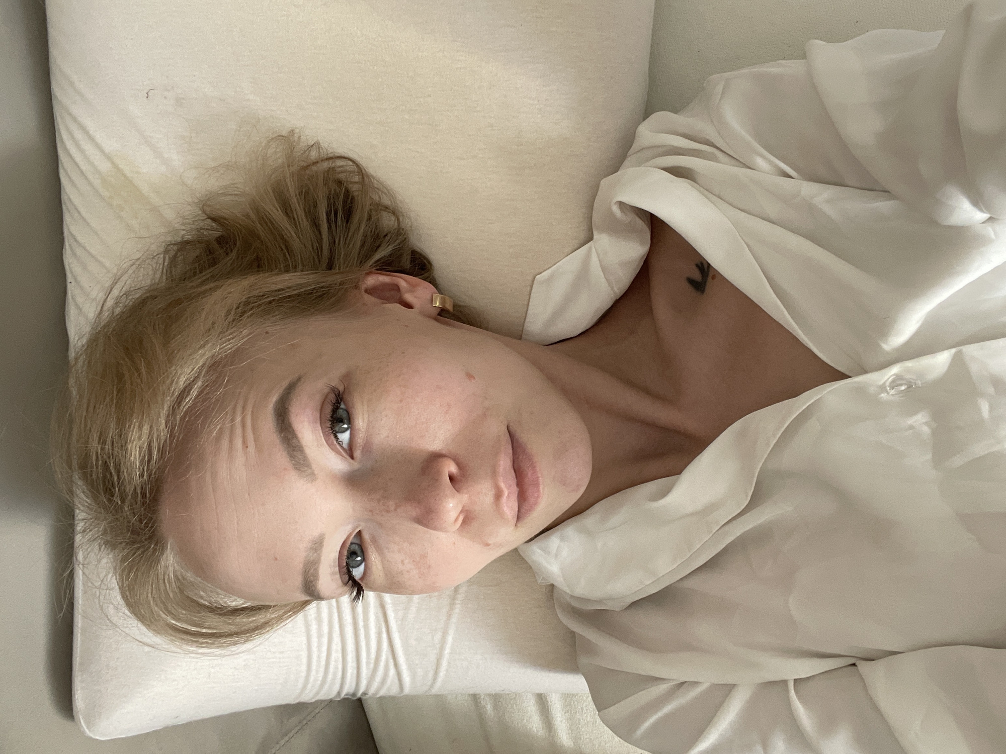 A blonde woman lies on a white pillow and is partially covered with a white shirt or sheet. She has freckles on her face and is looking at the camera. There is a tattoo on her upper chest. The location is unknown.