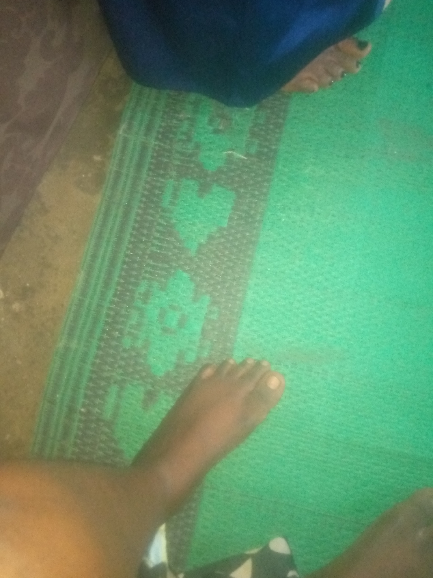The image shows a person's legs and feet on a green mat. The mat has a black and grey patterned border with floral designs. The person's feet are bare, and one foot has dark nail polish on its toenails. The background is blurred and appears to be a floor with some texture. There is a dark blue fabric partially visible in the upper portion of the image.