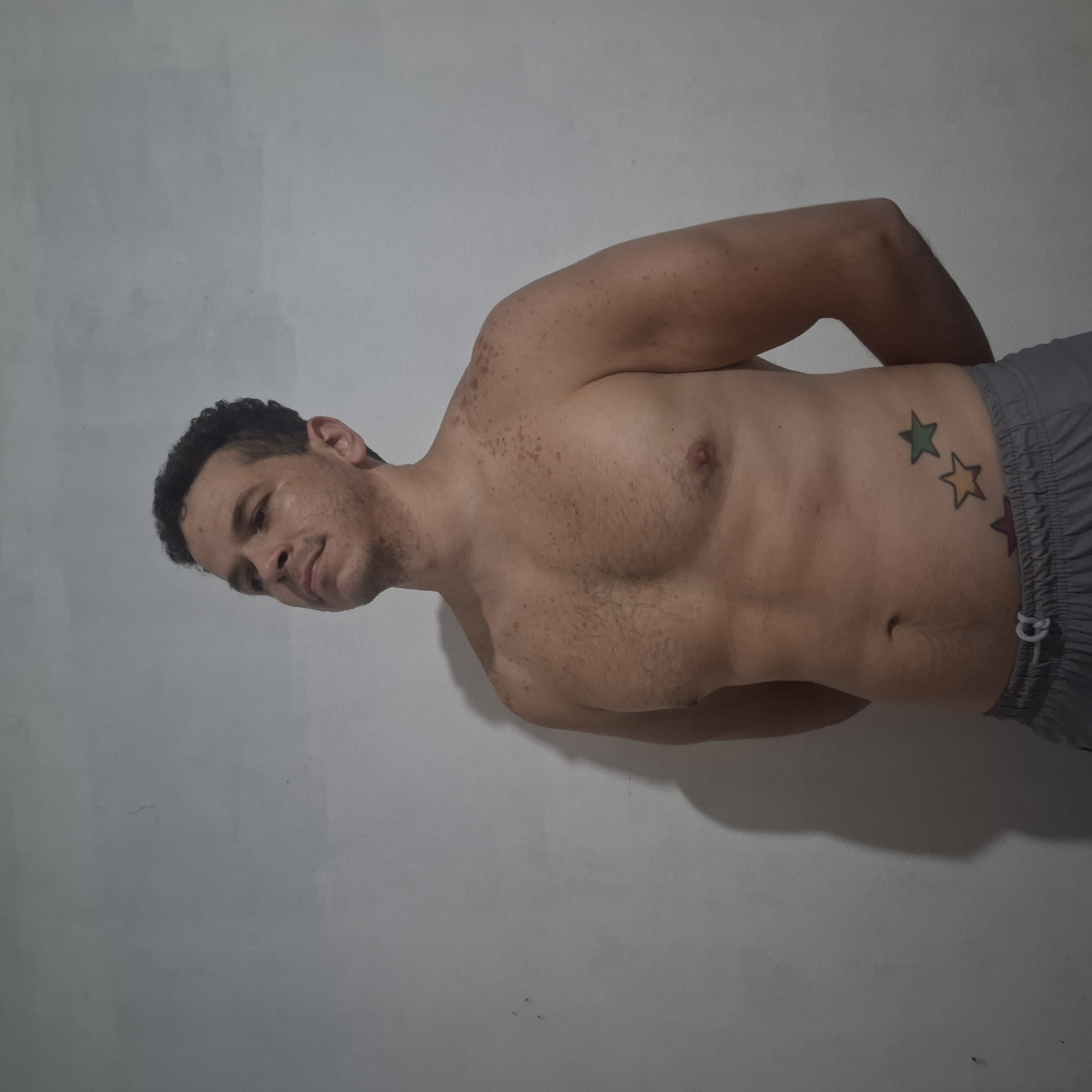 An adult male stands shirtless against a light-grey wall in what appears to be an indoor setting in Wilde, Argentina. He has short, dark, curly hair, stubble, and numerous small, dark brown macules distributed across his chest, shoulders, and neck. His arms are positioned behind his back, obscured from view. He is wearing grey athletic shorts with a white drawstring. On his left lower abdomen/hip, there is a tattoo depicting three outlined stars in an ascending diagonal arrangement: a reddish-brown star, a yellowish-orange star, and a green star. The man is looking upwards and slightly to his right. A soft shadow of his left side is cast onto the wall behind him.