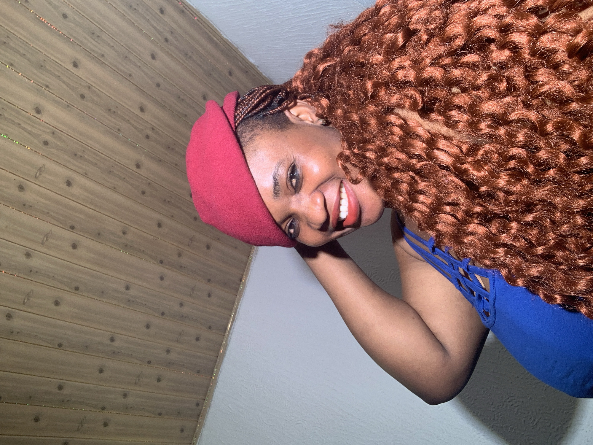 The image shows a close-up indoor portrait of a person, likely female, with dark skin, seen from the chest up. The person is smiling broadly, revealing white teeth and wearing light red lipstick. They have voluminous, reddish-brown curly hair extensions that cascade over one shoulder. Small braids are visible under a dark red beret worn on the head. The person is wearing a royal blue top with intricate, criss-cross strap detailing visible on the upper side.
The background is divided diagonally. To the upper left, there is a wooden panel wall or ceiling with distinct horizontal planks and dark circular knots. Interspersed between many of the wooden planks are thin, reflective, glittery lines that catch the light in various hues like orange, green, and gold. The lower right portion of the background is a plain, light-colored, subtly textured wall, possibly white or light grey. The person's right arm is bent, and the hand is out of frame. The overall expression is cheerful and friendly.
The image appears to be a selfie or a casual photo, captured indoors in Barakin Akawo, Nigeria, under artificial lighting conditions. There is no visible text.