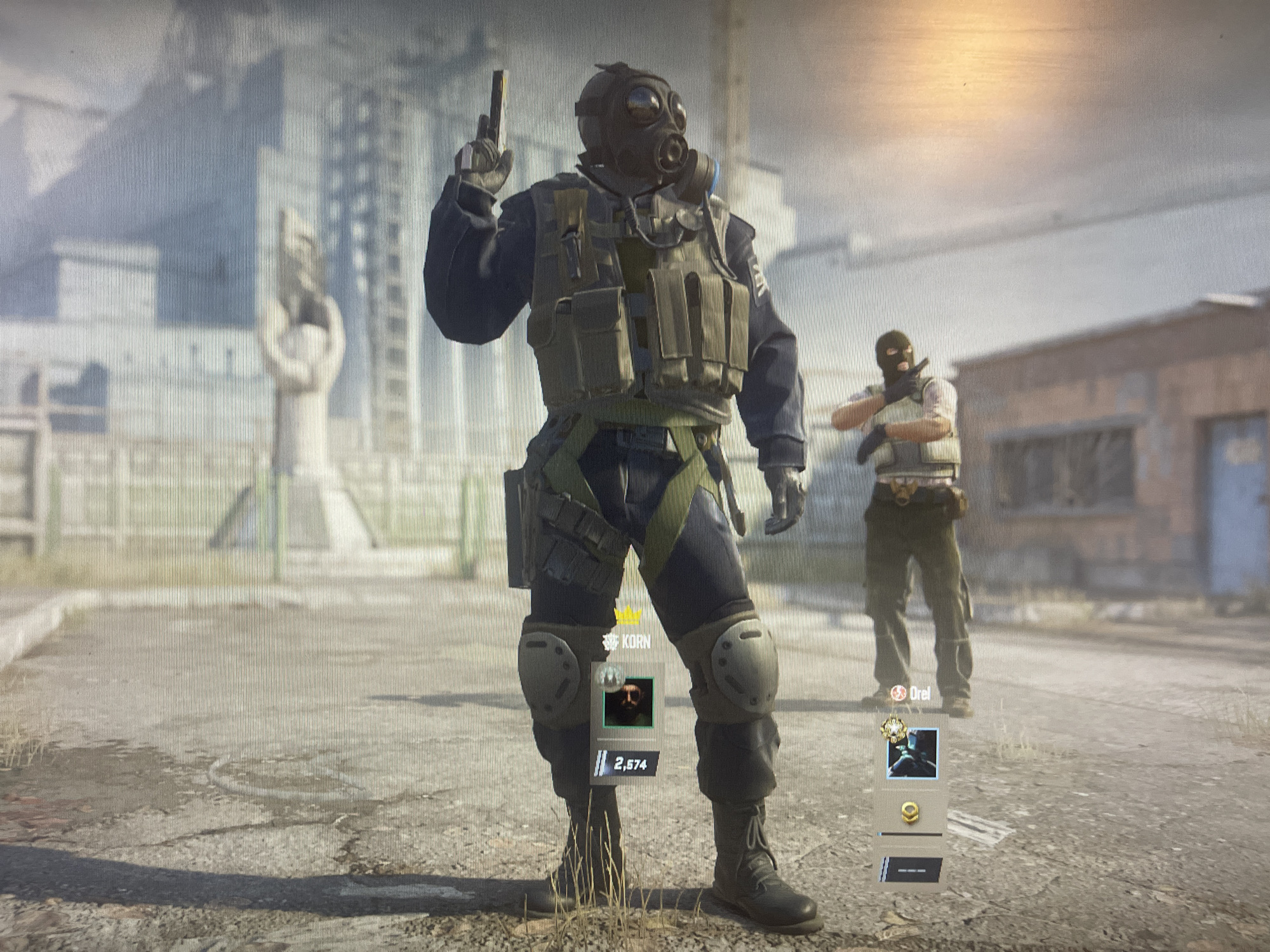 In an urban environment, two individuals are depicted in what appears to be a video game character selection or lobby screen. The foreground features a heavily equipped character wearing a gas mask and holding a pistol. This character has a vest with multiple pouches, knee pads, and combat boots. A player name "KORN" and a score of "2,574" are visible beneath them. To the right, a second character, wearing a black balaclava and a similar vest, stands with a pistol raised. This character's name is "Drel." The background suggests a post-apocalyptic or industrial setting with weathered buildings and debris. The lighting indicates an overcast or dimly lit day.