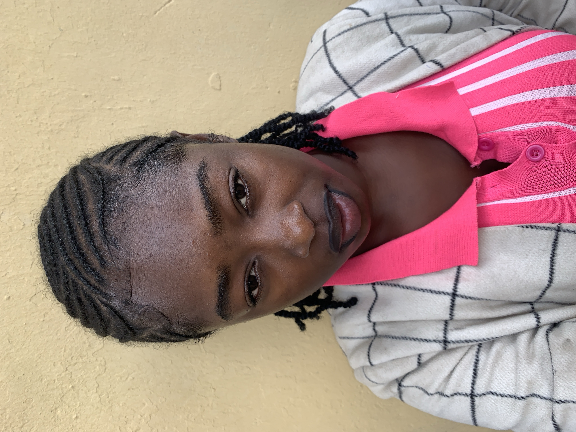 A close-up, head-and-shoulders portrait captured in Rafin Par, Nigeria, features an individual with dark skin, looking directly into the camera with a neutral expression. The subject's hair is styled in neat cornrows, with visible baby hairs along the temples. Her complexion appears dewy, and her lips are full, with a darker pigmentation in the center. She wears a bright pink polo shirt with white vertical stripes and a collar, fastened with two pink buttons. Over the pink shirt, she layers an off-white or cream-colored cardigan or jacket with a black grid pattern. The background consists of a uniformly lit, textured wall in a light yellow or beige hue.