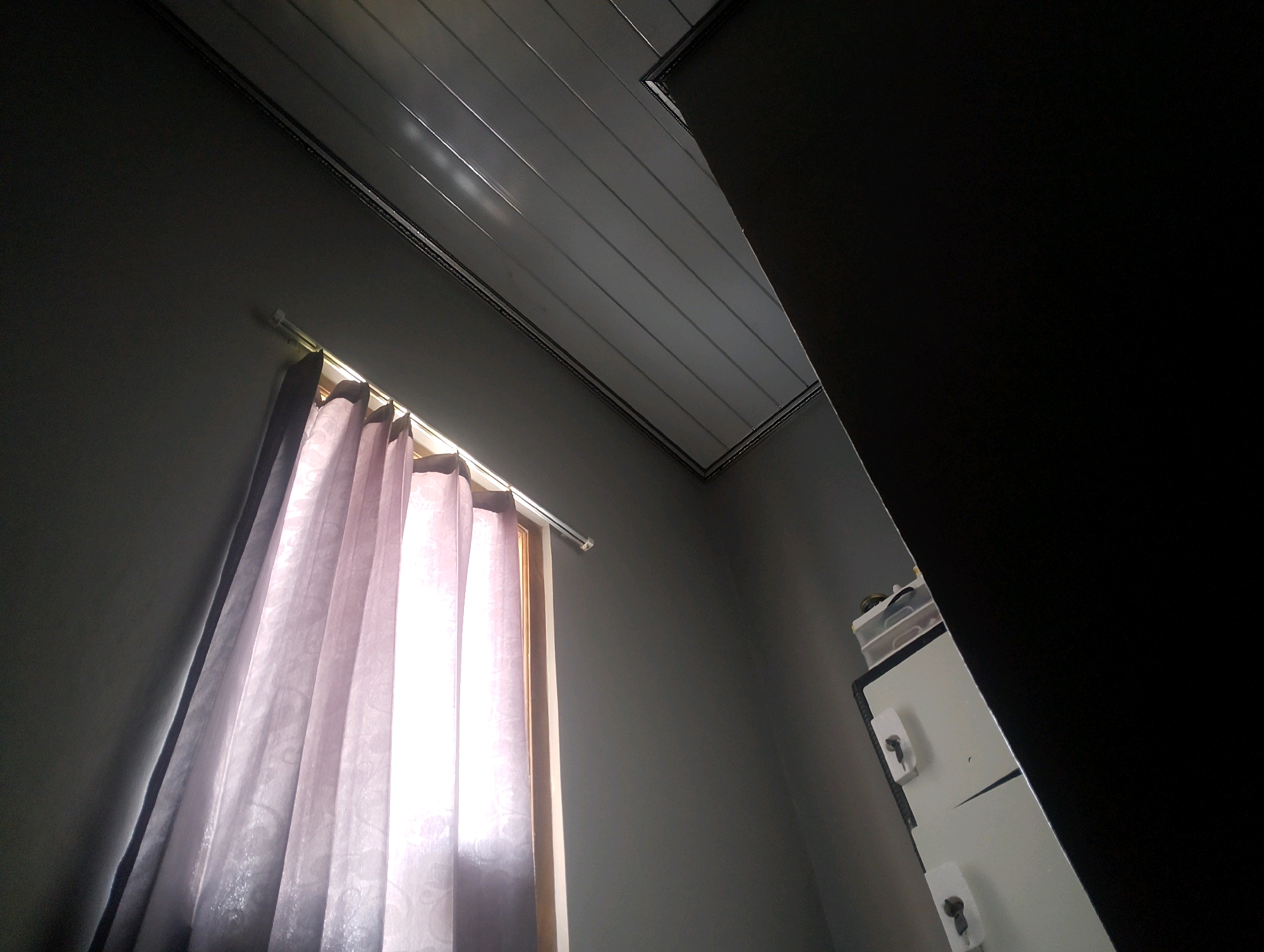 The image shows an interior scene from a low angle, looking upwards. The dominant elements are a section of a grey wall on the left, a light pink curtain hanging from a curtain rod, a window, and a dark, angular shape on the right that appears to be a piece of furniture or a doorway. Above, a white, paneled ceiling with horizontal lines is visible, bordered by dark trim.
On the right side of the image, partially obscured by the dark shape, is a piece of white furniture with visible drawers and knobs, suggesting it could be a cabinet or dresser. A small container with items is placed on top of this furniture.
The light source appears to be from the window behind the curtain, causing the curtain to be translucent and bright. The scene is indoors and does not provide any specific indicators of a location within Magelang, Indonesia, beyond being an interior space. There are no people, animals, or specific actions depicted. The overall impression is a dimly lit room with a window and some furniture.