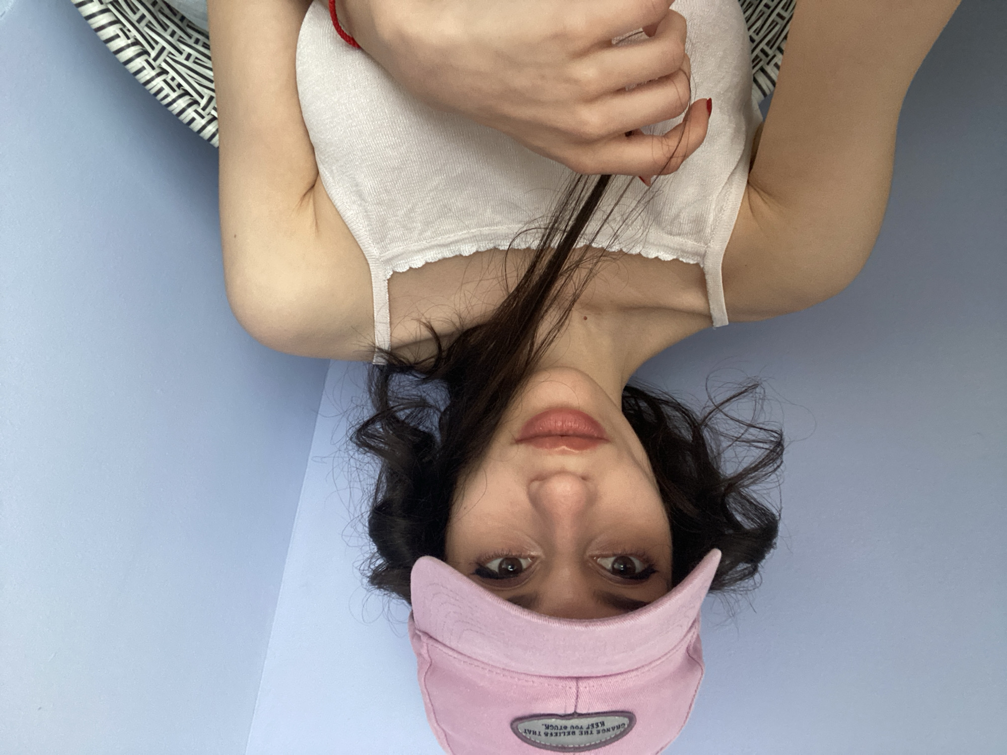 The media file is a close-up, upside-down selfie of a young woman. She has dark, wavy hair, brown eyes, and is wearing a white tank top and a pink baseball cap. A red string bracelet is visible on her left wrist. The woman appears to be lying down, as evidenced by the angle of her body and the fabric of a striped pillow or cushion visible in the upper left portion of the frame. The background is a solid, light purple-blue color. The lighting suggests it could be daytime, possibly indoors, given the even illumination. There is no indication of weather or a specific outdoor location. The woman's expression is neutral, and the overall mood is casual and relaxed. A small, oval patch is visible on the pink cap with text that reads, "CHANGE THE BELIEFS THAT KEEP YOU STUCK."