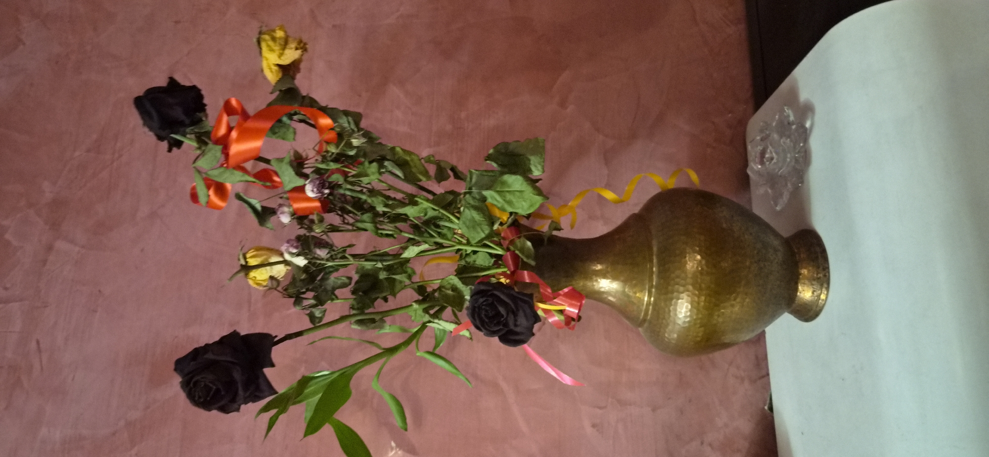 The image displays a still life composition featuring a bouquet of flowers arranged in a metal vase. The vase, positioned towards the center-right, is crafted from a hammered, brass-colored metal, exhibiting a warm, golden hue and a textured surface.
The bouquet contains a variety of roses and foliage. Several prominent black roses are visible, alongside yellow roses that appear to be in various stages of drying or wilting. Smaller, withered roses with faded purple or pinkish tones are also present. Lush green leaves and stems form the base of the arrangement, some stems being long and slender. Brightly colored ribbons are interwoven among the flowers; these include vibrant orange satin ribbons, thin yellow curly ribbons, and a delicate pink ribbon, adding decorative accents.
The vase and flowers are placed on a flat, white surface, likely a table or shelf, which extends across the right side of the frame. On this white surface, near the top right, a small, clear glass object shaped like a star or flower is visible. The background consists of a textured wall painted in a reddish-brown or terracotta shade, showing subtle variations in color and finish. The lighting suggests an indoor setting. There are no people or active events depicted, and no visible text.