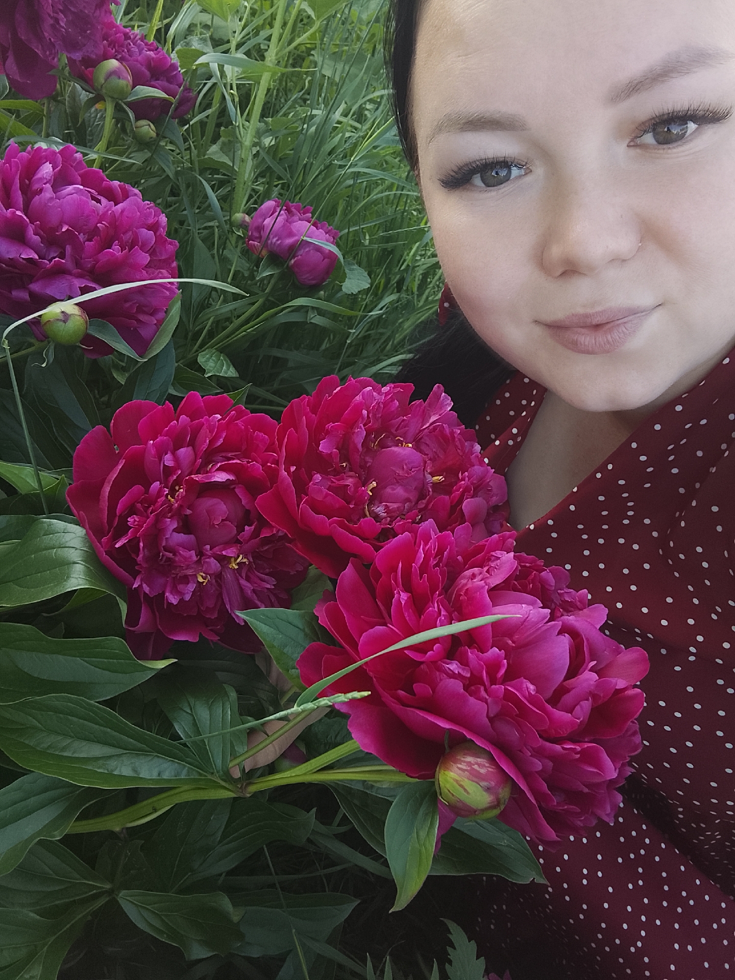 A young woman with dark hair and prominent eyelashes takes a selfie outdoors, surrounded by lush green foliage and vibrant magenta peonies. She is wearing a red dress with white polka dots. The background appears to be a garden or natural setting. The lighting suggests it is daytime, possibly late afternoon given the soft quality of light. The image's composition places the woman's face in the upper right quadrant, with the peonies dominating the left and bottom portions of the frame. The overall scene is peaceful and evokes a sense of summer. The presence of peonies suggests this scene is taking place during late spring or early summer, the blooming season for these flowers.