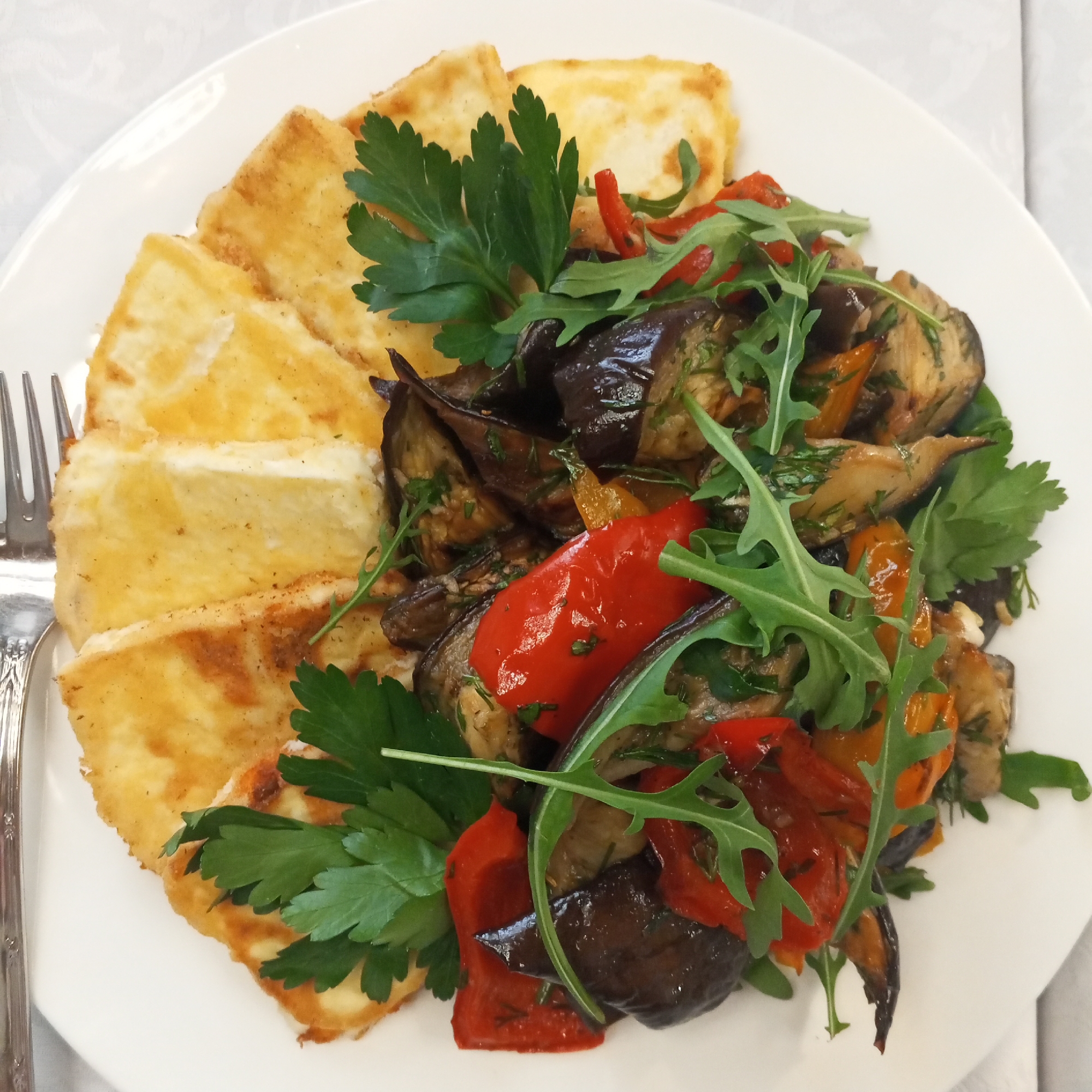 A top-down shot shows a white plate with a meal. The plate contains fried halloumi cheese, sauteed vegetables (eggplant, red and yellow bell peppers), parsley, and arugula. A fork is visible on the left side of the plate. The meal rests on a table covered with a white tablecloth that has a subtle, textured pattern. The image was taken in Chaikovsky, Russia.