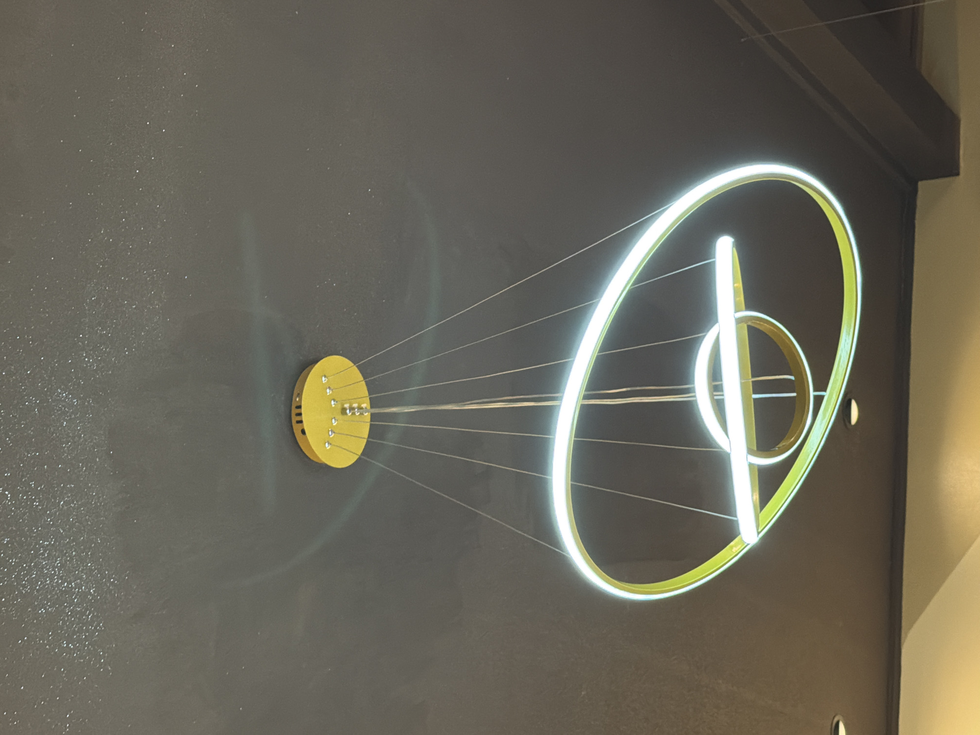 A modern, abstract chandelier is mounted on a textured, gray wall. The chandelier features a yellow circular base from which thin wires extend to a large, glowing, elliptical ring. Within the ring are a smaller gold circle and a vertical white bar. The scene is indoors, with the upper right corner showing a dark gray ceiling and a light-colored wall in the background. This image was taken in the city of Kuta, Indonesia.
