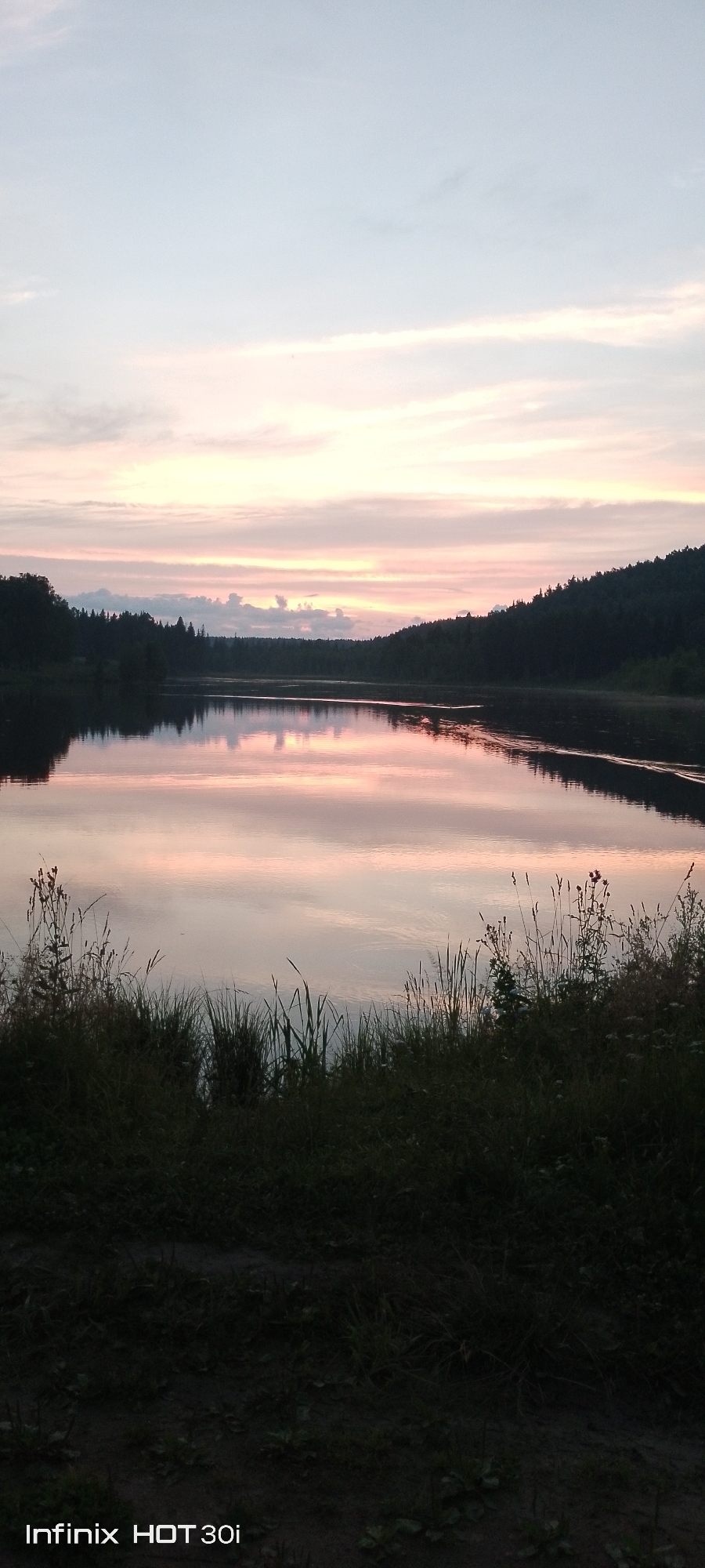 This is a vertical, outdoor, full-shot photograph of a serene lake scene at dusk. The sky above is a soft gradient of pale blue at the top transitioning to delicate pink and orange hues near the horizon, indicative of sunset or sunrise. Wispy clouds are scattered across the sky, catching the light.

The lake in the midground is calm and reflective, mirroring the colors of the sky and the dark silhouettes of the trees that line its banks. The trees form a dense, dark band across the horizon, with a few taller evergreens standing out. The shoreline in the foreground is covered in tall, wild grasses and some flowering plants, which are silhouetted against the lighter sky and water. The overall mood of the image is peaceful and tranquil.

The time of day is clearly dusk, with the soft, diffused light and the colors in the sky. The weather appears to be clear and calm, as evidenced by the smooth surface of the lake. The setting is natural and undeveloped, suggesting a rural or wilderness location.

Visible text at the bottom left corner reads: "Infinix HOT 30i".