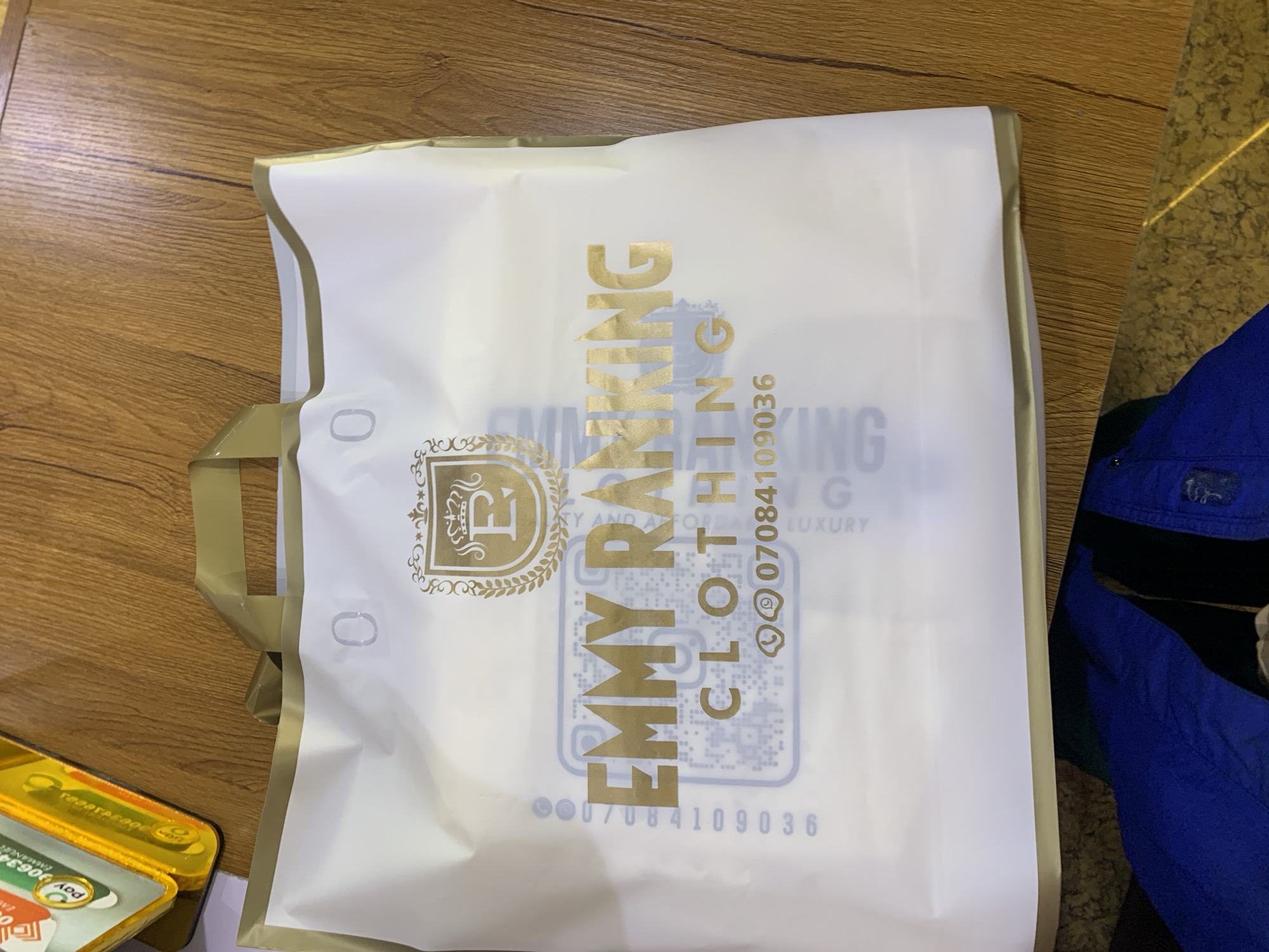 A white shopping bag with gold lettering and a gold emblem is placed on a wooden surface. The shopping bag displays the words "EMMY RANKING CLOTHING" in gold lettering, along with a phone number "07084109036" and a QR code. In the upper left corner of the image, a yellow and green package is partially visible. The background appears to be a wooden floor. The shopping bag and its contents are the primary subjects of the image, with the wooden surface and the partial package serving as environmental context. No people or actions are depicted. The image is a still photograph of these objects.