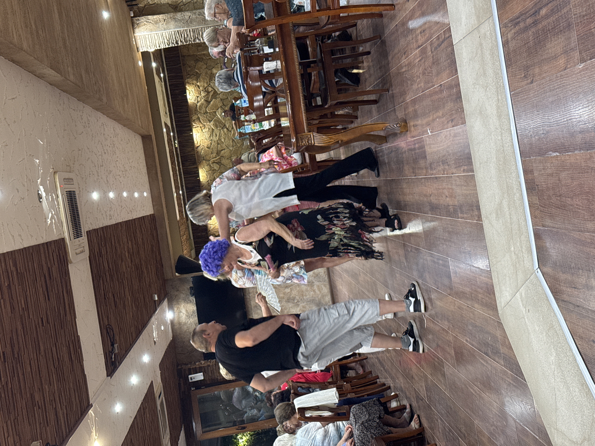 The image depicts a group of people inside what appears to be a restaurant or a banquet hall. The setting features a wooden floor and ceiling with decorative elements. In the foreground, a man in a black t-shirt and grey shorts is interacting with a woman wearing a vibrant purple wig and a floral dress. Another woman in a white top and black pants is also visible. Several other individuals are seated at tables in the background, some of whom are eating or conversing. The ambiance suggests a social gathering or event. The location is identified as Sredets, Bulgaria.