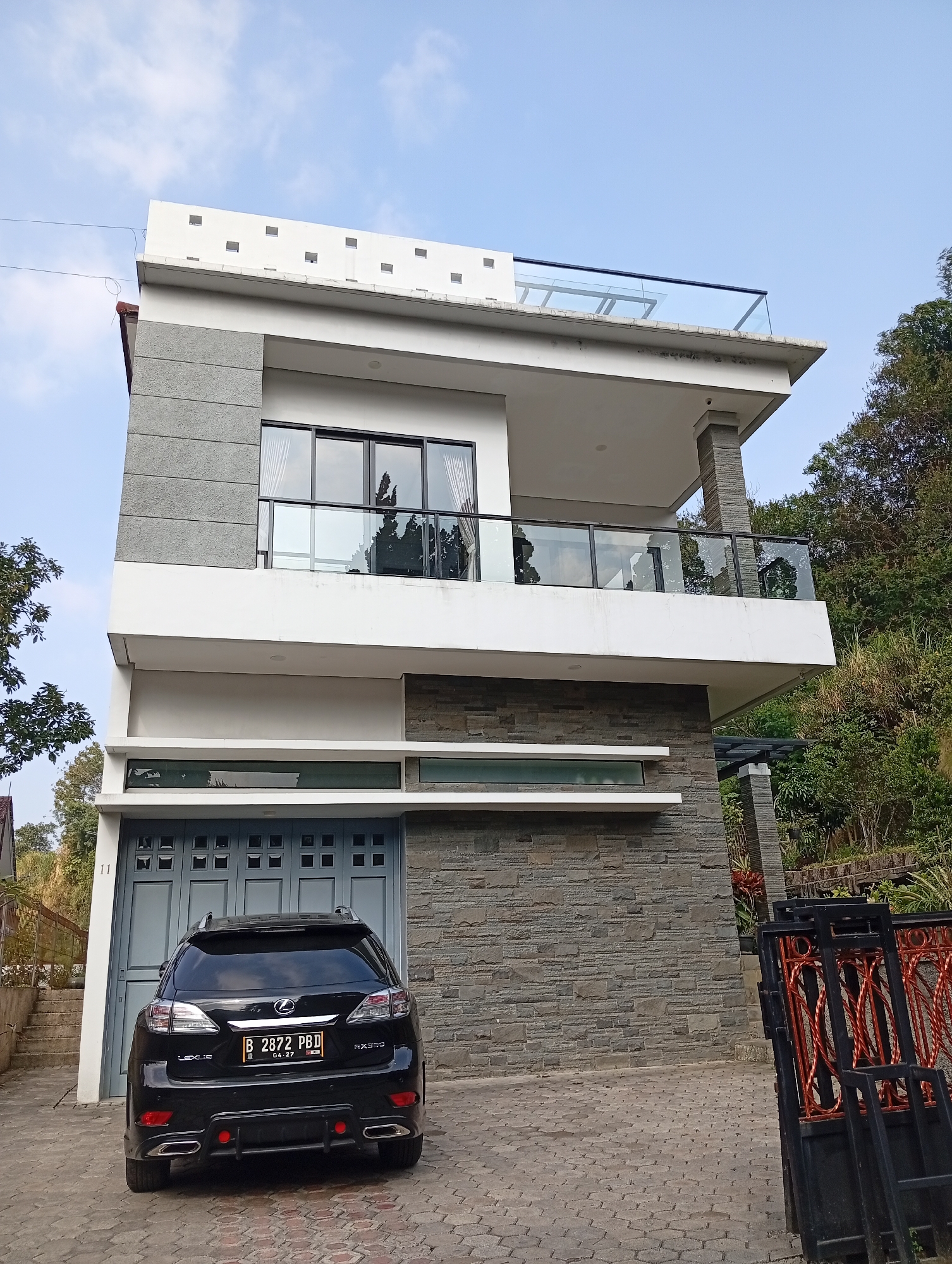 The image depicts a modern, multi-story house located in Cimacan, Indonesia, situated on a sloped terrain with lush green trees and vegetation. The house features a contemporary architectural style with clean lines, a combination of white, gray stone, and concrete finishes. It has a prominent glass-enclosed balcony on the upper level and a large garage door on the ground floor. A black Lexus RX350 with a license plate reading "B 2872 PBD" is parked in front of the garage. Adjacent to the property is a black metal gate with decorative red scrollwork. The sky above is a clear blue with scattered clouds.
