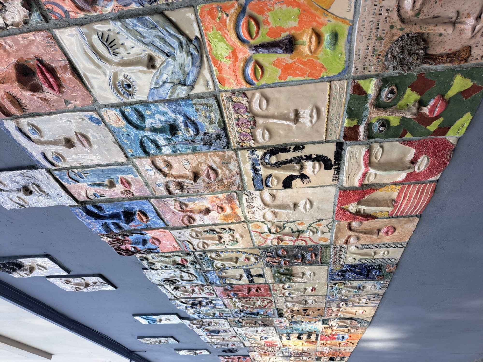 The image captures a unique artistic installation, likely adorning a ceiling. It comprises numerous square tiles, each featuring a sculpted human face with diverse expressions and colors. The tiles are arranged in a dense grid pattern, creating a visually striking mosaic of faces.
This artwork is located in Yerevan, Armenia, though the specific venue is not provided in the prompt or visible in the picture.
The style of the faces varies, with some tiles displaying abstract or surreal interpretations, while others appear more realistic. The color palette is similarly diverse, ranging from muted earth tones to vibrant primary colors. The overall effect is a thought-provoking and somewhat unsettling exploration of human identity and expression.