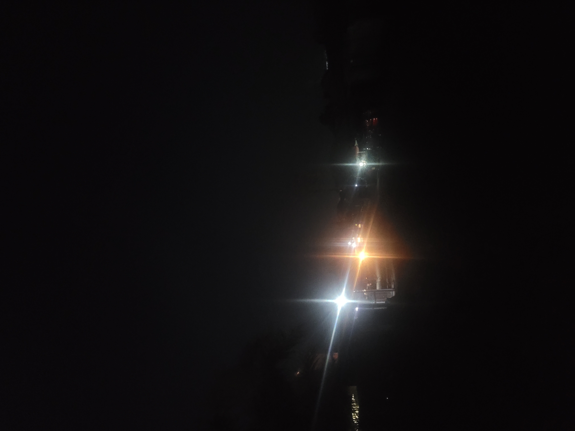 The image displays a predominantly dark, nighttime scene with a vertical band of bright artificial lights on the right side. The majority of the frame, especially the left and upper areas, is deep black, indicating very low ambient light.
The subjects are primarily sources of intense light and their reflections. Several strong light sources are visible, producing significant lens flares that create starburst effects. One light in the upper right is bright white, while another below it exhibits a warm, yellowish-orange glow. These lights appear to be mounted on or associated with a structure, possibly a bridge, an elevated road, or a building. Faint details of what might be metallic railings or structural elements are subtly visible around the illuminated areas.
Below the main light sources, clear reflections of the lights are present in what appears to be a calm body of water, suggesting the scene is situated near a river, lake, or another large water expanse. The reflections stretch downwards, mimicking the light patterns above.
The activity depicted is static, a snapshot of an illuminated structure at night. The time of day is clearly night, with no natural light. There are no discernible people or visible text in the image. The extreme contrast between the dark surroundings and the brilliant artificial lights is the most notable detail, creating a dramatic visual. Given the location context, this scene is from Jos, Nigeria.