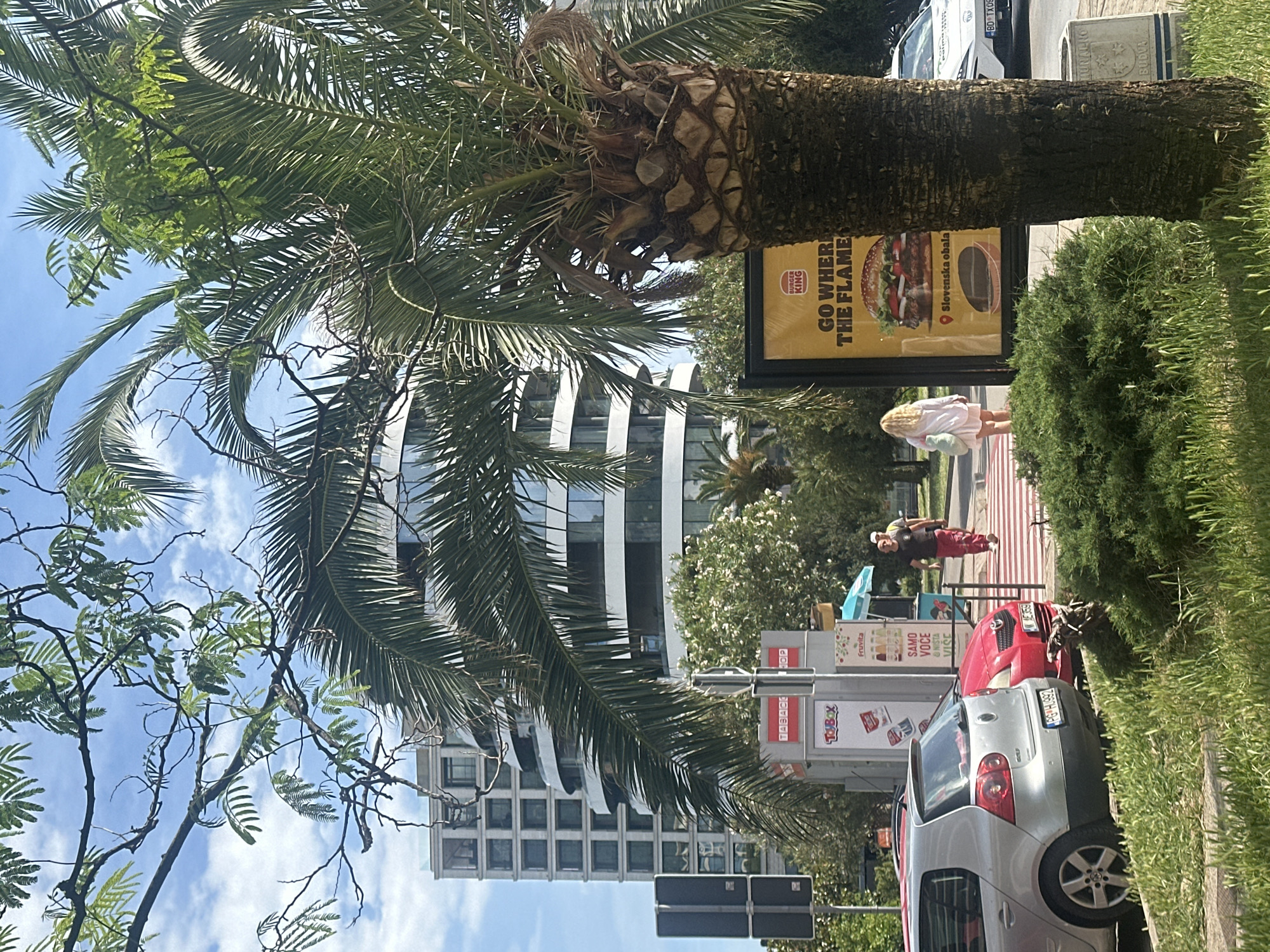 The image depicts a street scene in Budva, Montenegro. A Burger King advertisement showing a hamburger is visible on a billboard near a palm tree. A woman in a white dress is walking near the billboard. In the background, a modern building is partially obscured by the tree branches. Parked cars, including a silver Volkswagen and a red vehicle, line the street. Street signs and a bus stop are also visible. There are pedestrians on a crosswalk. The sky is blue with some clouds.