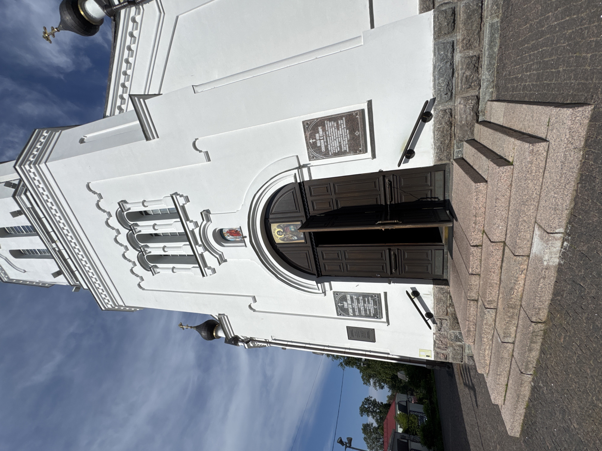 This image shows the exterior of a white church in Lomonosov, Russia. The church features dark wooden double doors, arched windows, and a black dome topped with a golden cross. Two plaques are affixed to the building's white facade. The left plaque, a dark rectangular stone, contains inscriptions in Cyrillic, including the dates "1896-1996" and the name "СЕРГЕЙ К. СИМОНОВ". The right plaque is also a dark rectangular stone with Cyrillic text.
A stone staircase leads up to the church entrance, with the steps made of rough-hewn pink granite. The surrounding ground is paved with dark cobblestones. The sky above is blue with scattered white clouds, suggesting a clear day. The sun is casting shadows, indicating it is likely daytime. The overall impression is one of a well-maintained, historical religious building.