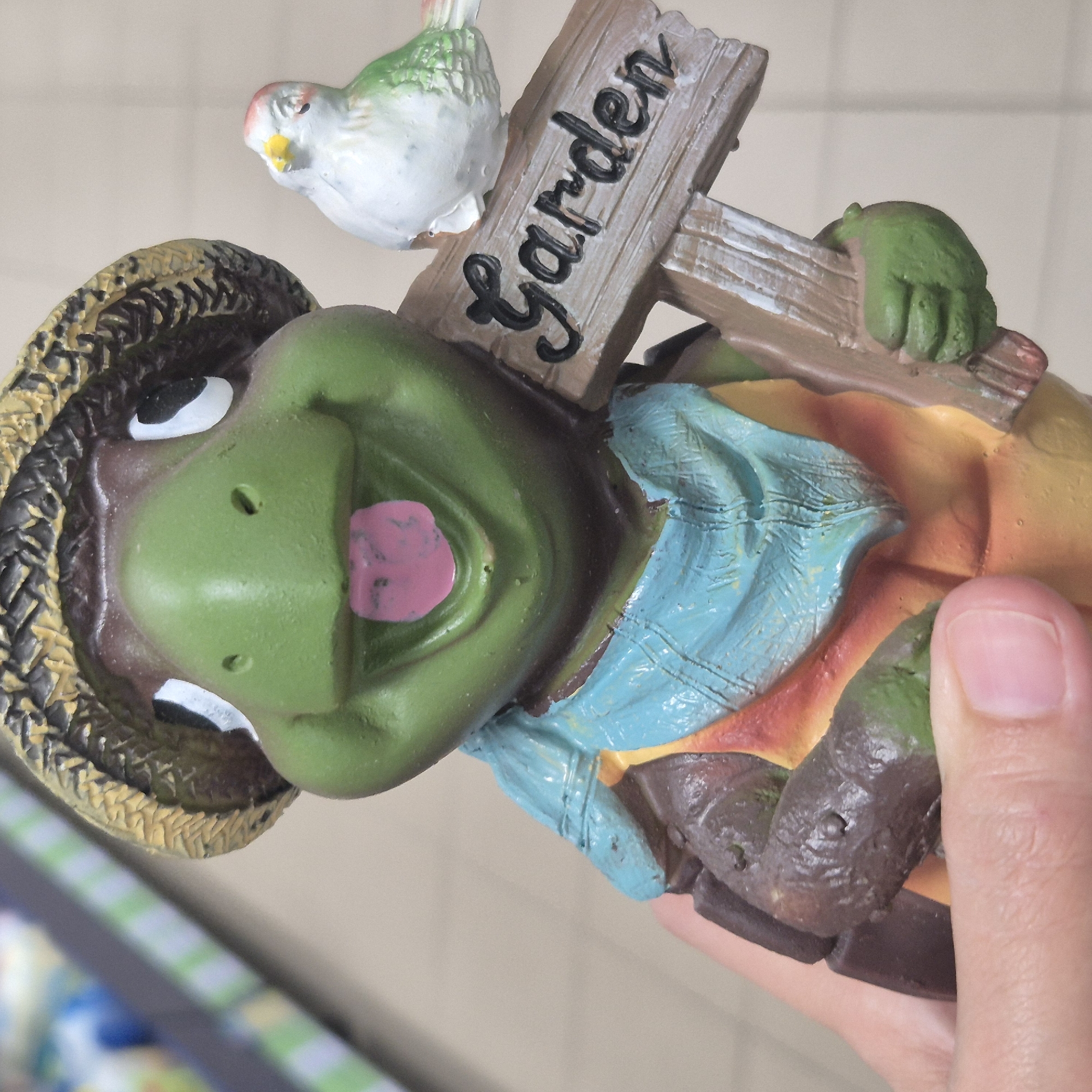 A close-up, slightly upward angled shot features a decorative garden statue of a smiling cartoon turtle wearing a straw hat and a light blue scarf. The turtle is holding a small wooden sign that reads "Garden" in black cursive. A small white and green bird is perched on top of the sign, facing left. The statue is held in a person's hand, with the thumb and index finger visible. The background is blurred and appears to be an indoor setting, possibly a store, with rows of products on shelves in the upper left corner. The overall impression is a whimsical and friendly garden decoration. The image was taken in Kokshetau, Kazakhstan.
