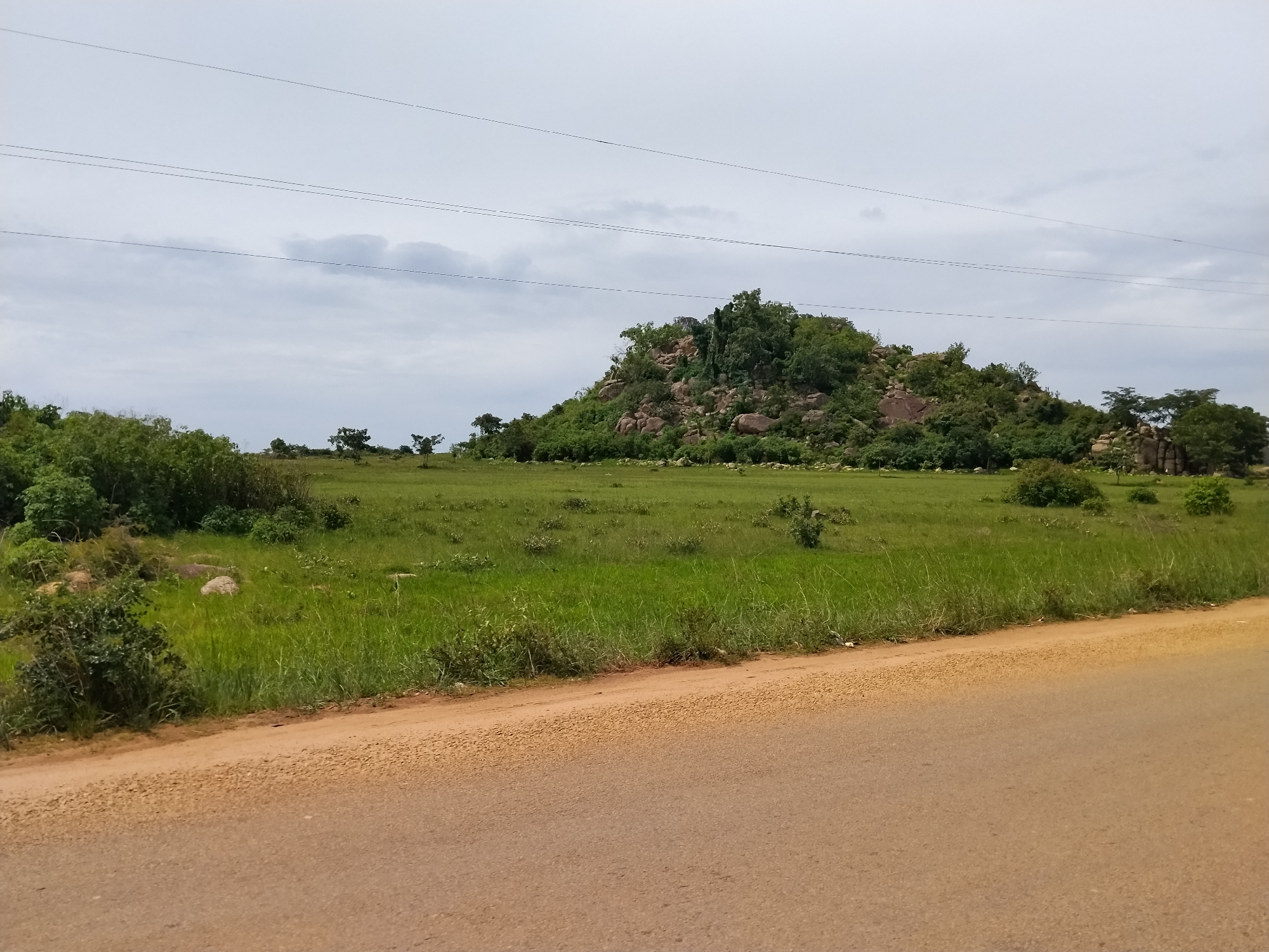 A wide-angle, eye-level, outdoor shot shows a landscape with a dirt road in the foreground. The road runs diagonally from the bottom left to the bottom right of the frame. Beyond the road is a large, grassy field dotted with shrubs and bushes. In the background, a rocky hill covered in trees rises from the field under a cloudy sky. Several power lines crisscross the sky. The image is presumed to be taken in Nabatum, Nigeria, based on its appearance.